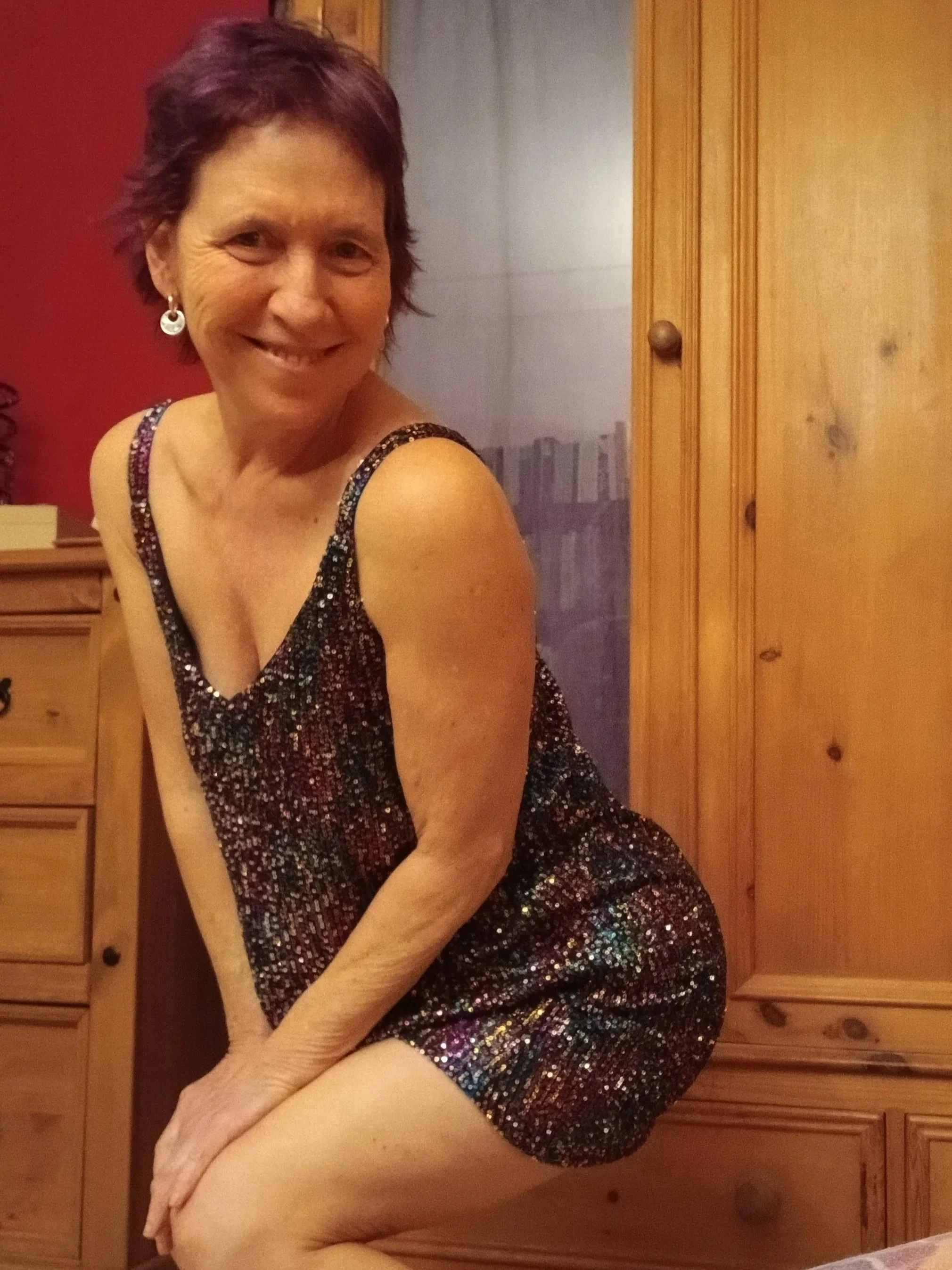 A smiling woman with short purple hair in a sparkle dress, leaning forward with hands on her knees in a room with wooden furniture.