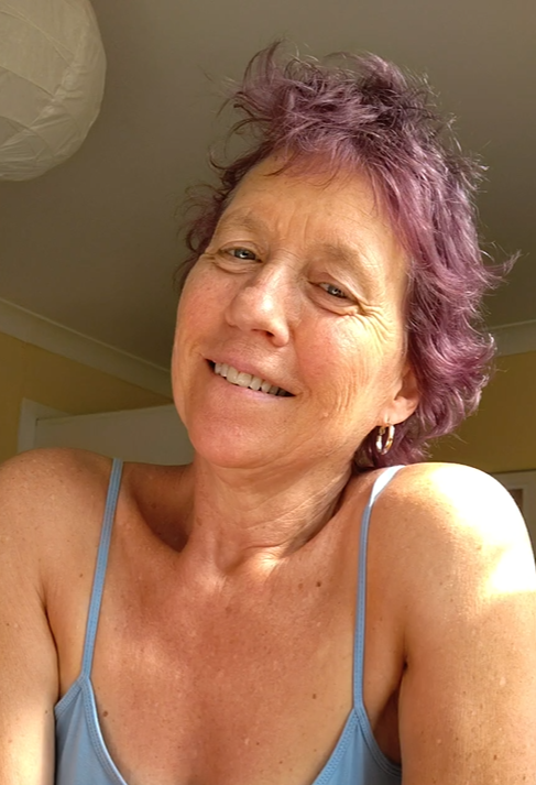 Maria Mountain - Intimacy and Connection Coach with short curly purple hair wearing a light blue spaghetti strap top, indoors.