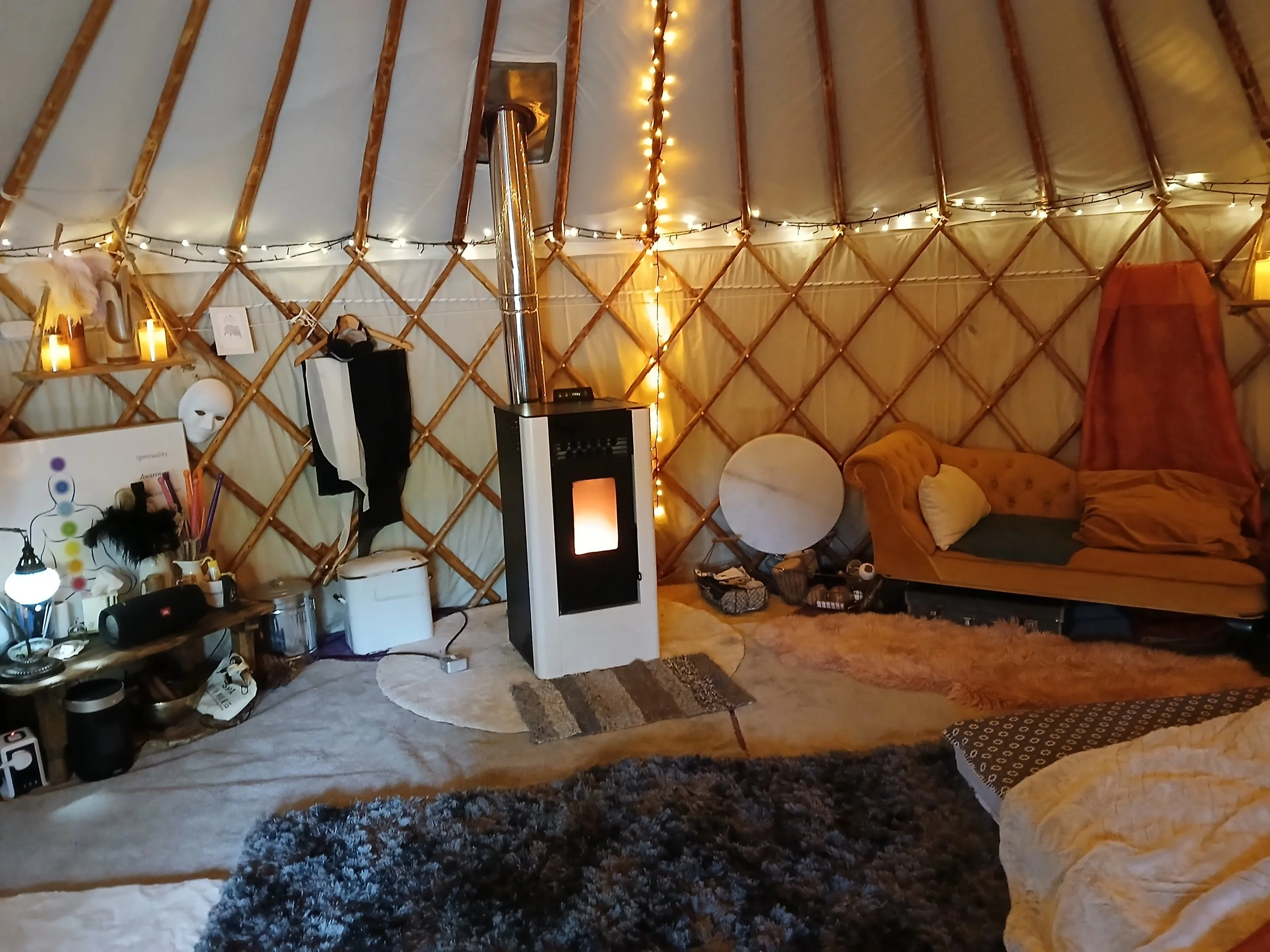 Interior of a cosy yurt with string lights, a wood stove, a yellow couch, and various decorations including candles, masks, and a small bookshelf.