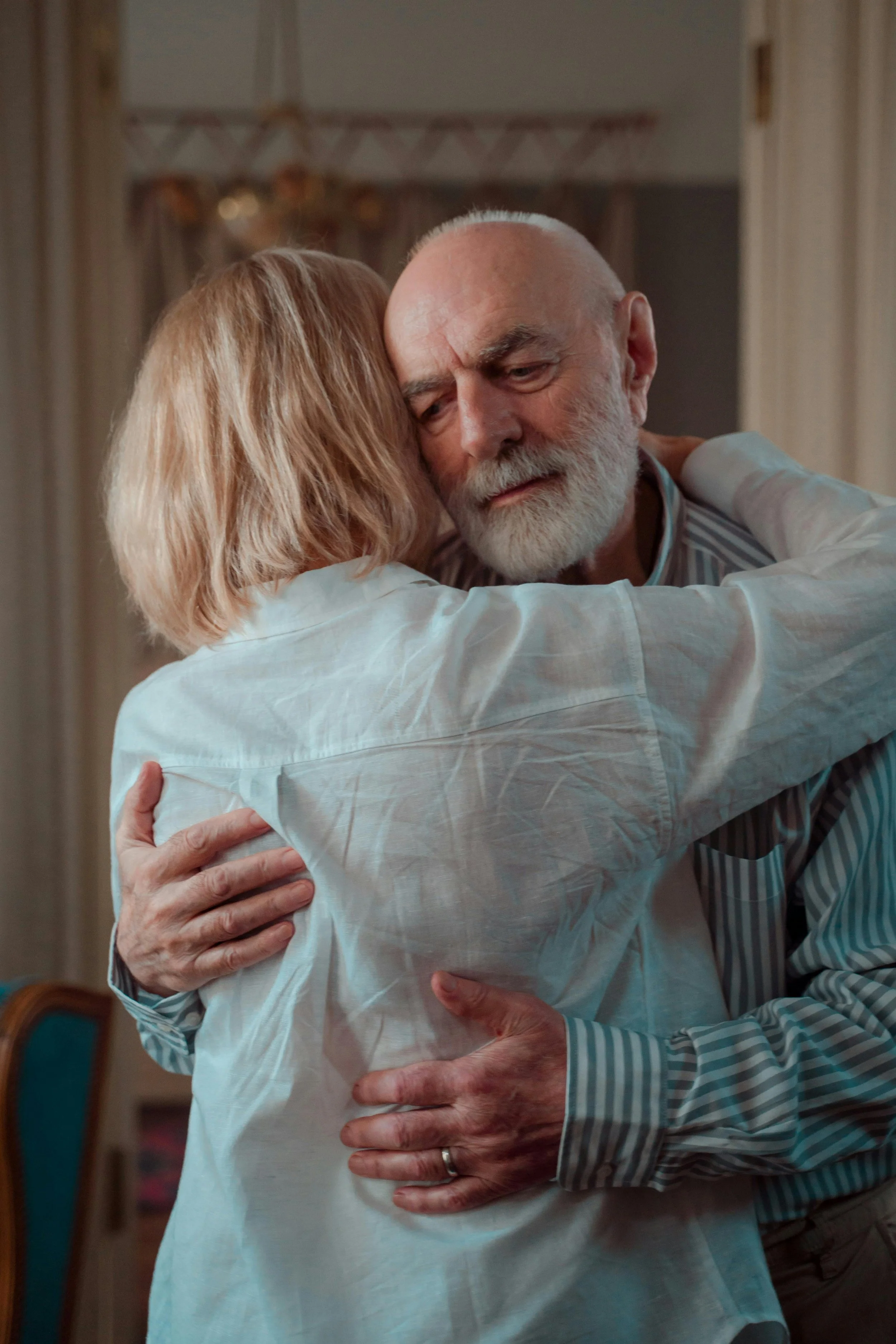 An elderly man with a white beard embraces a woman, both with their eyes closed, in a heartfelt hug indoors. Maria Mountain - Intimacy and Connection Coach