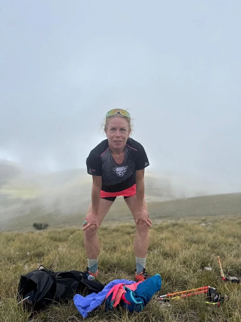 athletic woman with hands on knees on the top of a mountain, it's cloudy and her backpack is on the ground with her belongings unpacked.