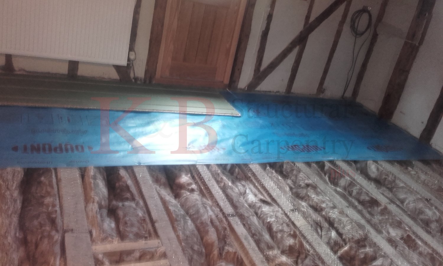 Construction in progress inside a wooden structure, with insulation materials on the floor and a sheet of metal leaning against the wall.