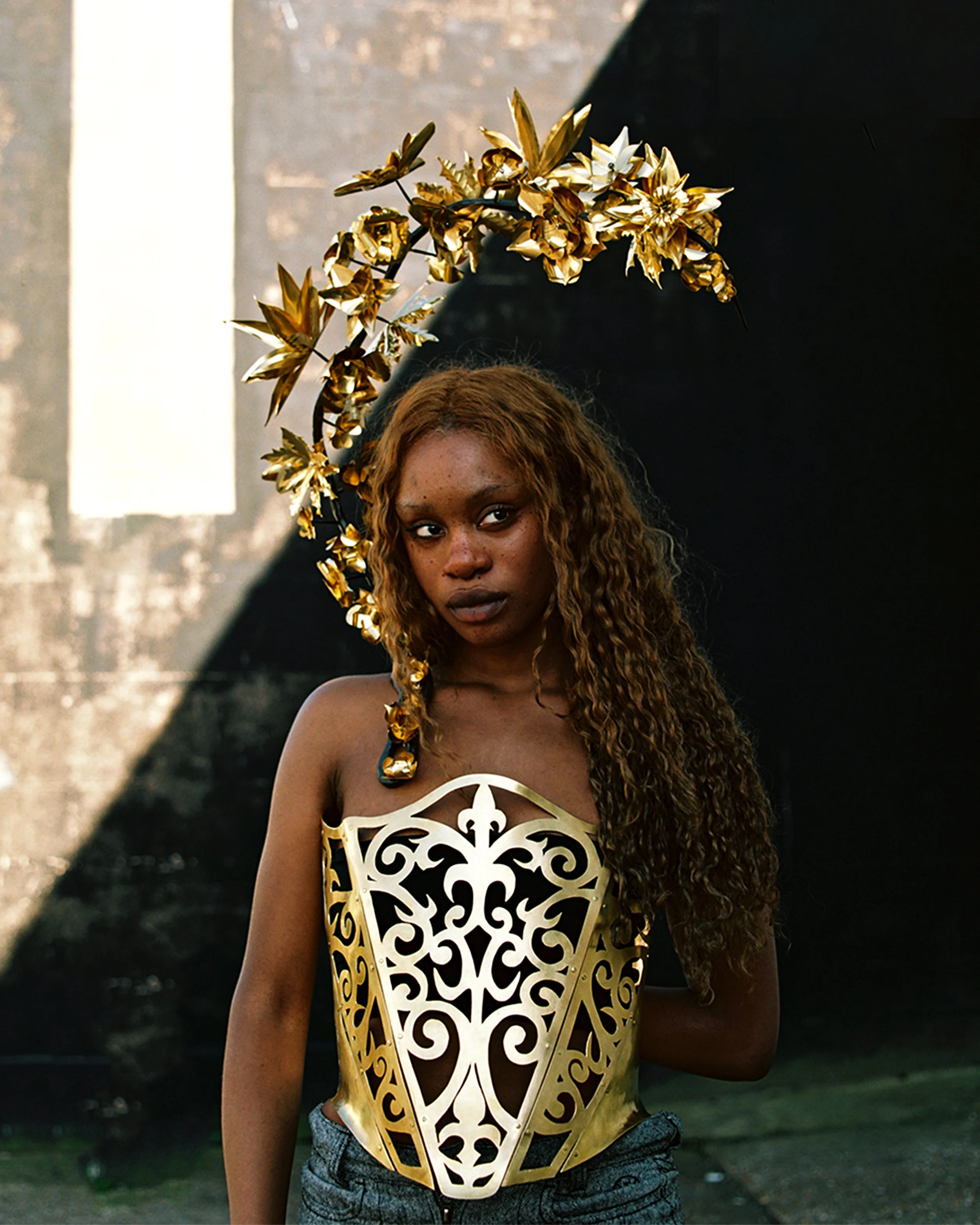 A woman with long curly hair wearing a gold decorative headpiece and a gold ornate top with open patterns, standing outdoors against a background of dark and light geometric shapes.