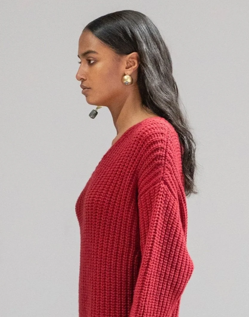 Side profile of a woman with long dark wavy hair, wearing a red knit sweater, and a headset microphone, standing against a plain gray background.
