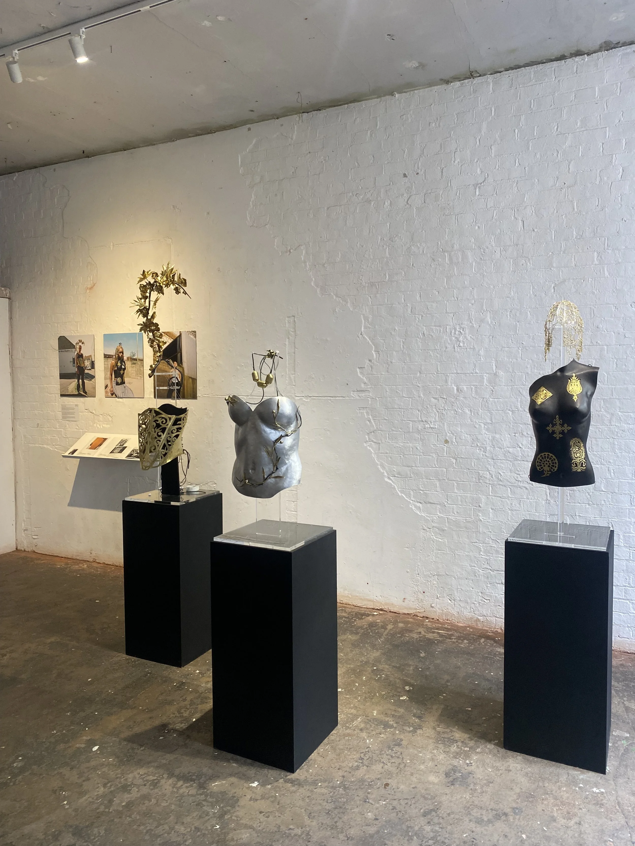 Art exhibit with three mannequin torso sculptures on black pedestals, decorated with metallic accessories and objects, against a white brick wall with photos and informational display.