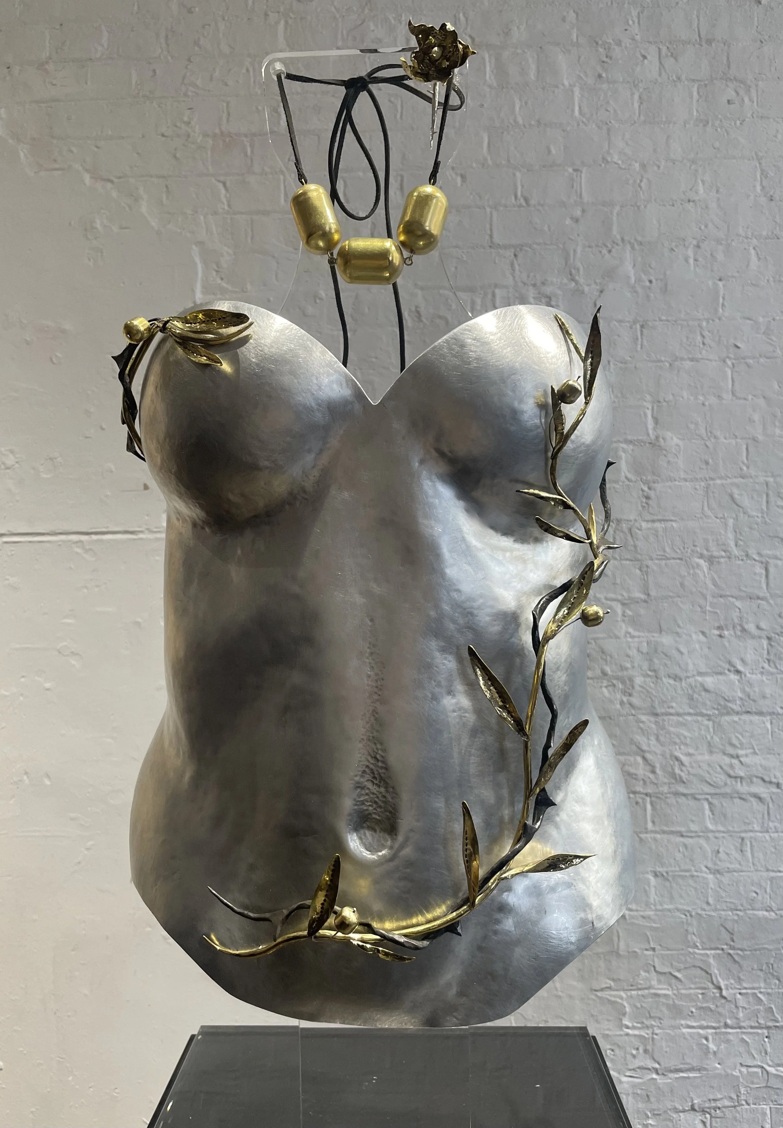 A metallic sculpture of a female torso with gold accents, decorated with gold leaves and beads, displayed against a white brick wall.