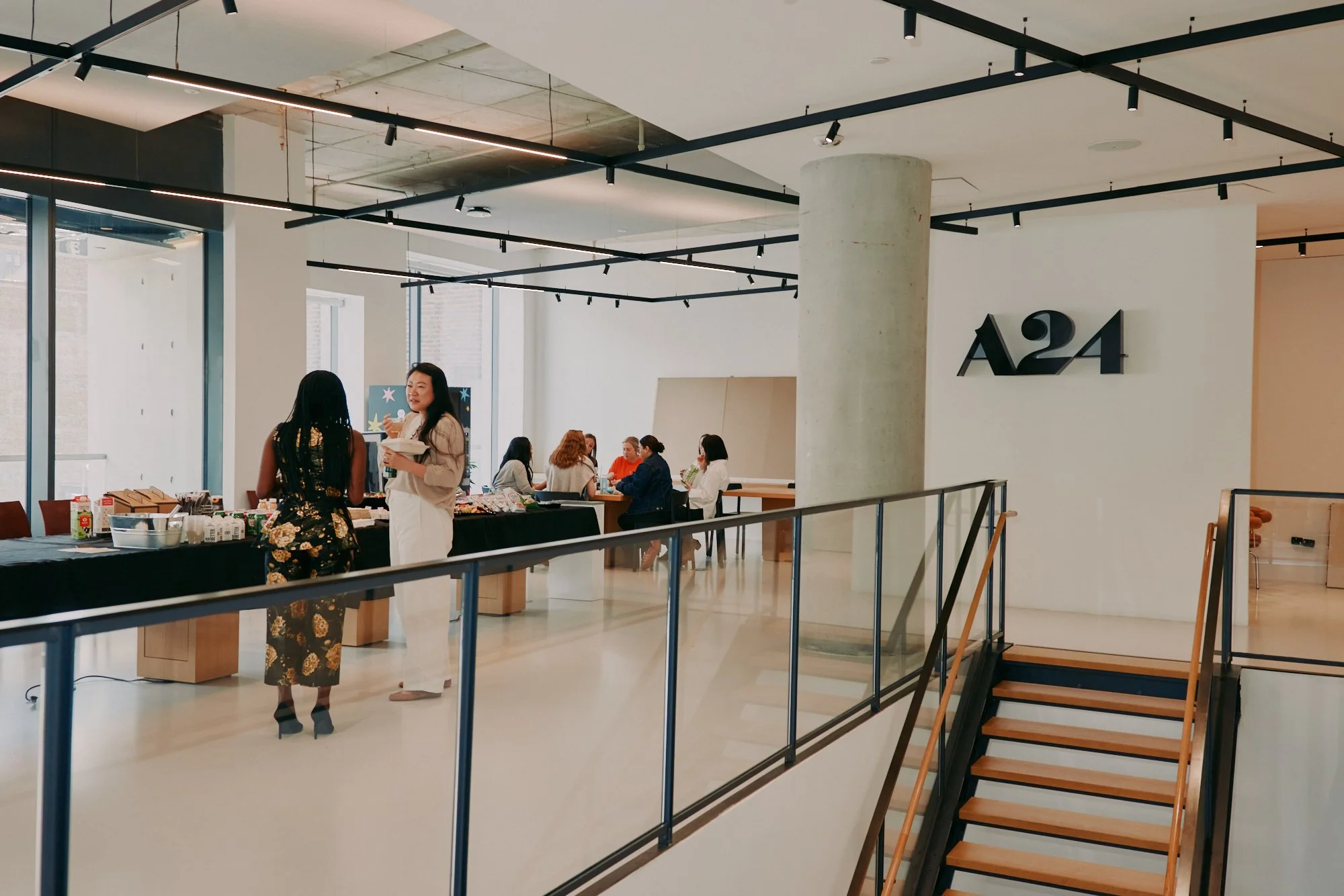 People socializing in an art gallery or showroom with wooden tables displaying items, large windows, and a sign that reads 'A24' on the wall.