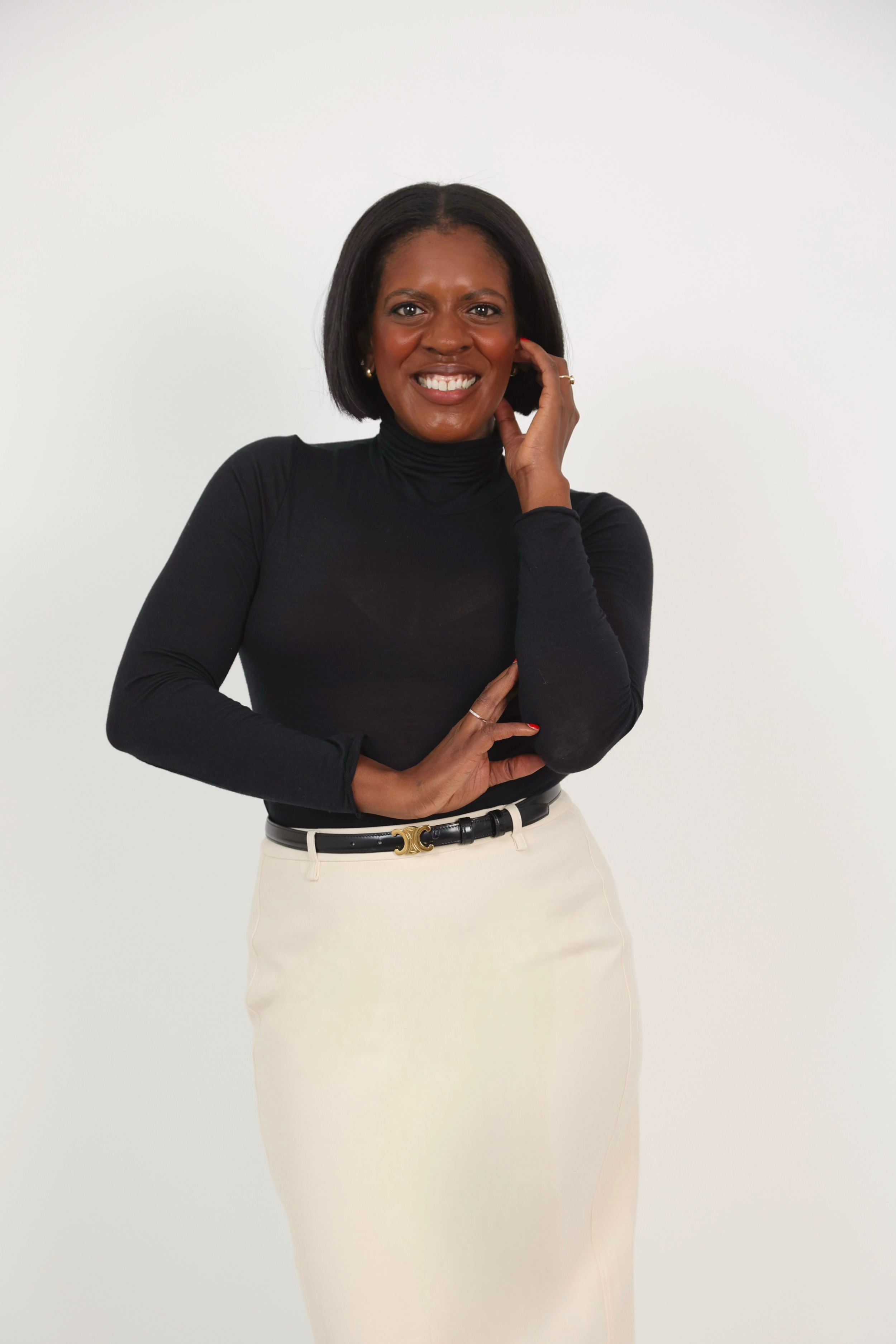 A woman with shoulder-length black hair wearing a black turtleneck and white skirt standing against a plain white background.