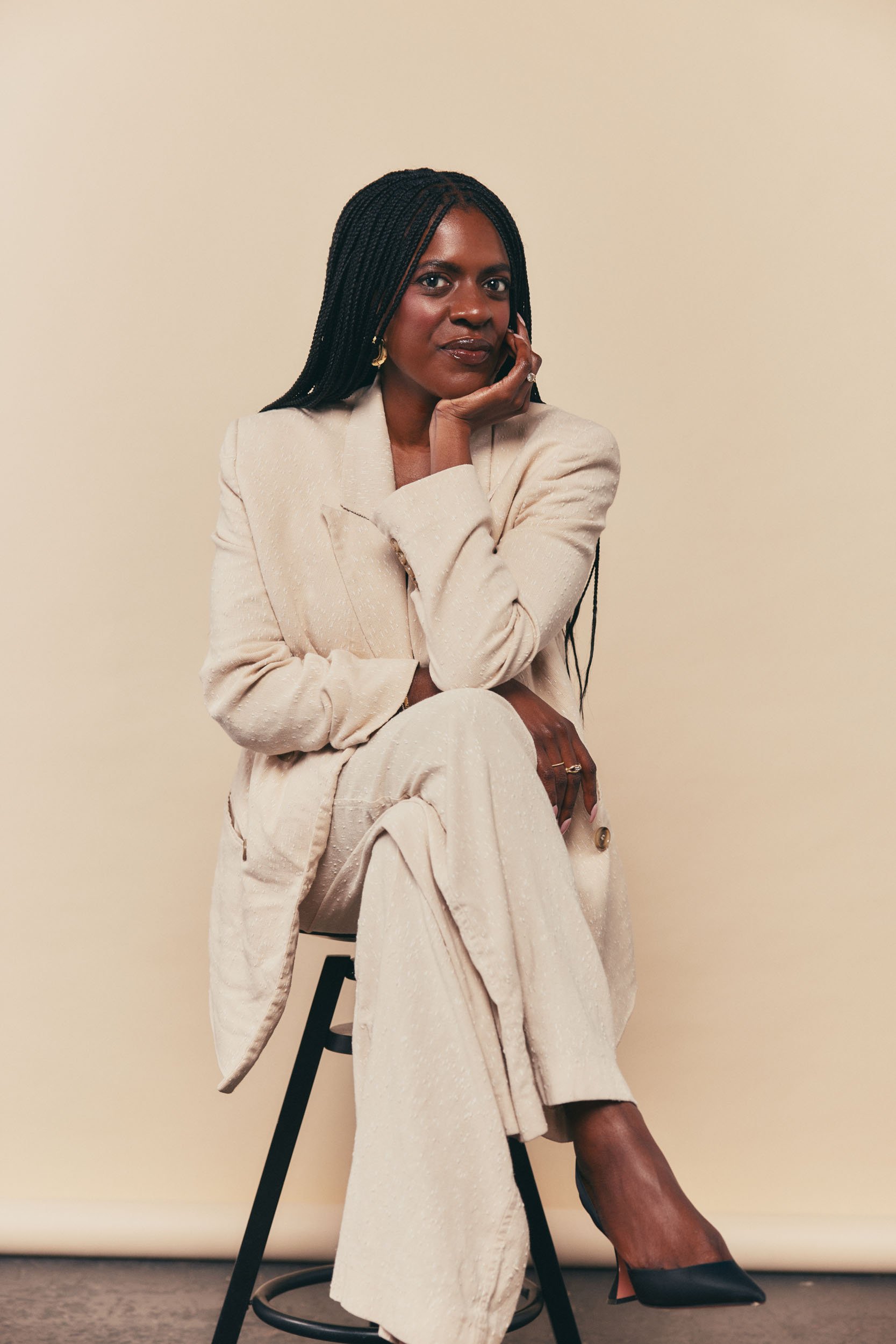 A woman with dark skin and long braided hair, wearing a beige blazer and matching pants, sitting on a black stool against a plain beige background, with her chin resting on her hand and looking directly at the camera.