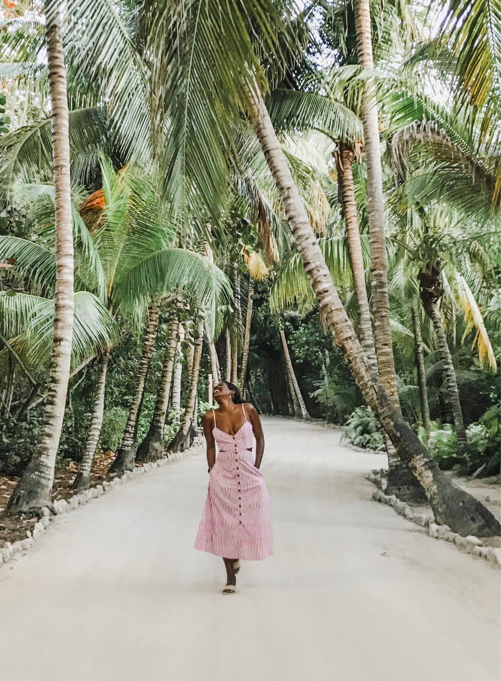 A woman in a pink and white striped dress walking on a sandy path through a lush tropical forest with tall palm trees.