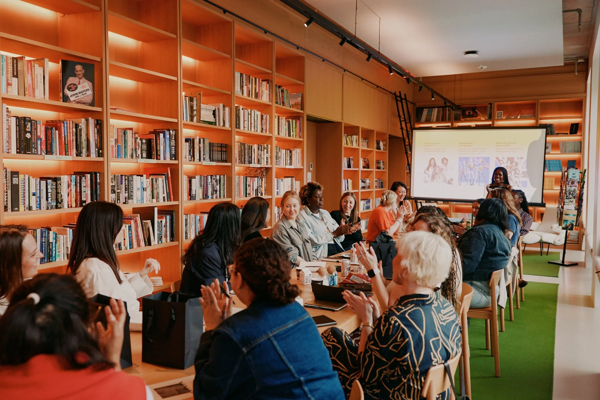 A group of diverse women sitting at a long table in a bookstore or library, attending a presentation or event. A woman is speaking near a projection screen, and the women are clapping and smiling.
