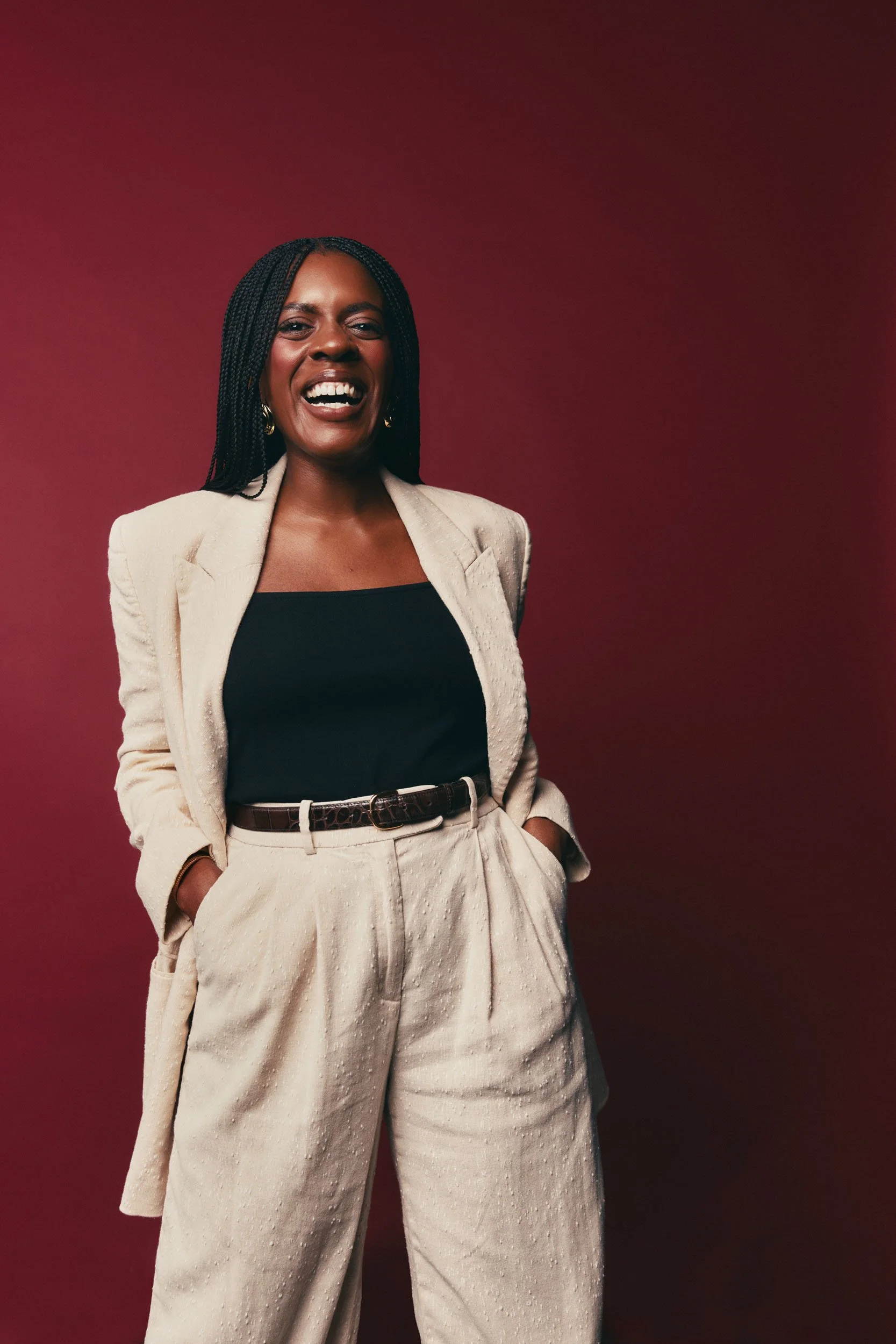Portrait of a smiling Black woman with braids, wearing a beige blazer and matching pants, black top, standing against a dark red background.