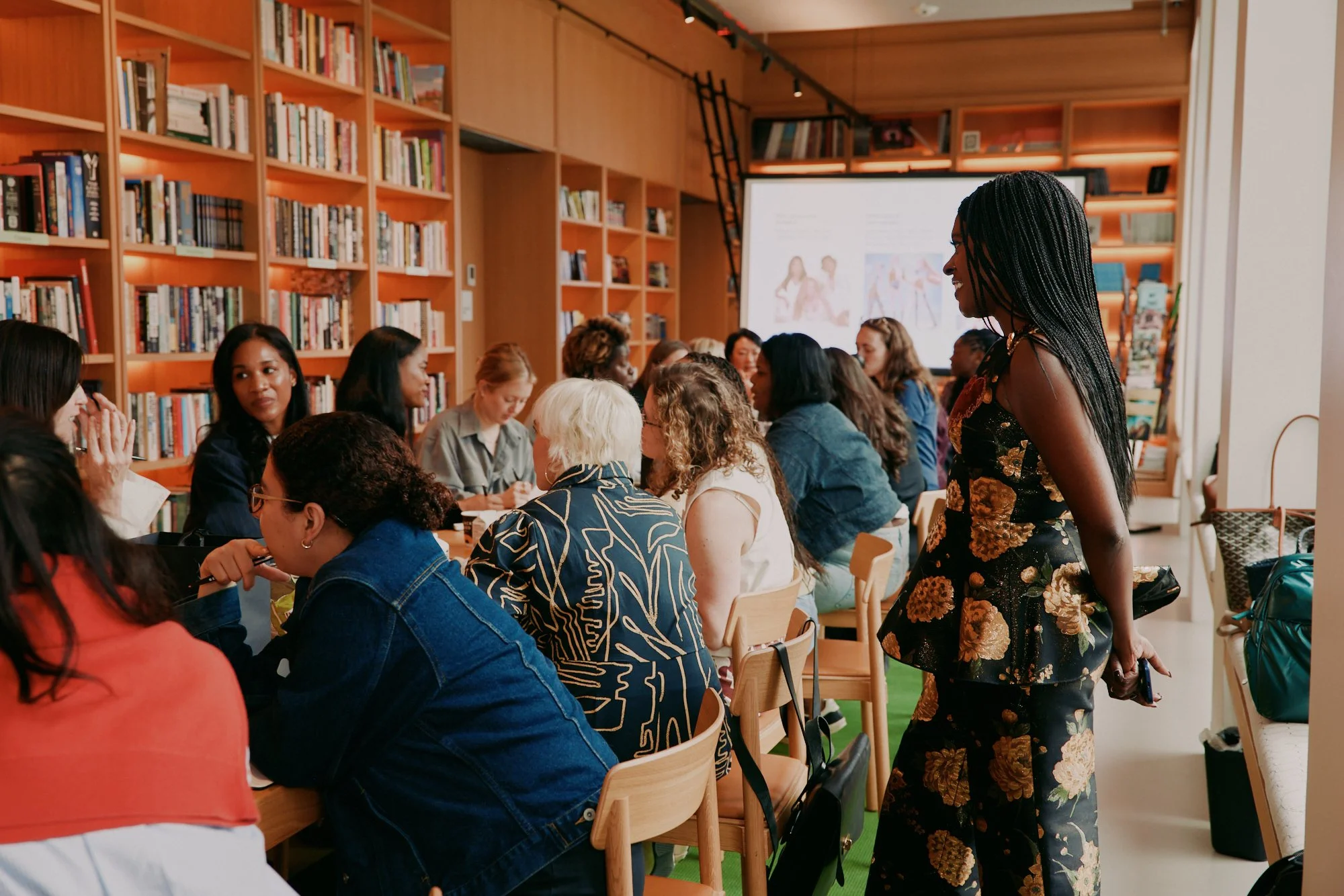 A diverse group of women sitting around a table in a room with wooden shelves filled with books. A woman with long braided hair stands nearby, smiling and engaging with others. There is a large screen in the background displaying images and text.