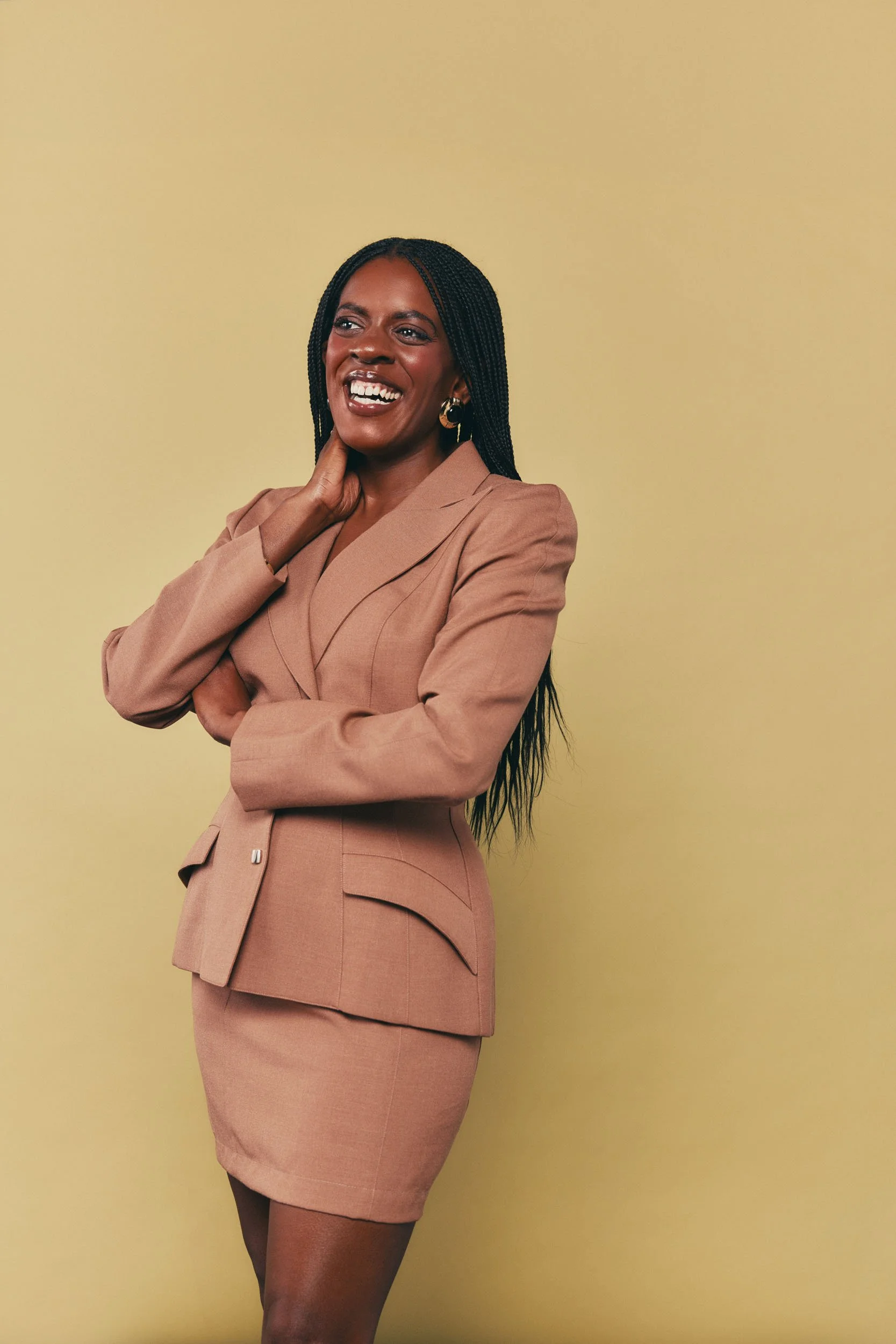 A confident woman with dark skin, long braided hair, and hoop earrings, wearing a beige blazer and matching pencil skirt, stands against a plain yellow background. She is smiling and has one hand touching her neck.