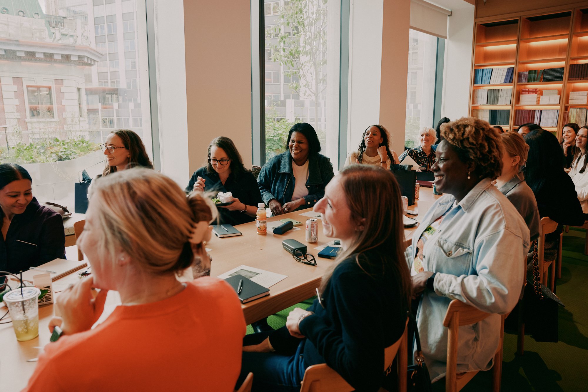 Group of diverse women gathered around a large conference table in a bright room with large windows and a bookshelf, engaging in a meeting or discussion.