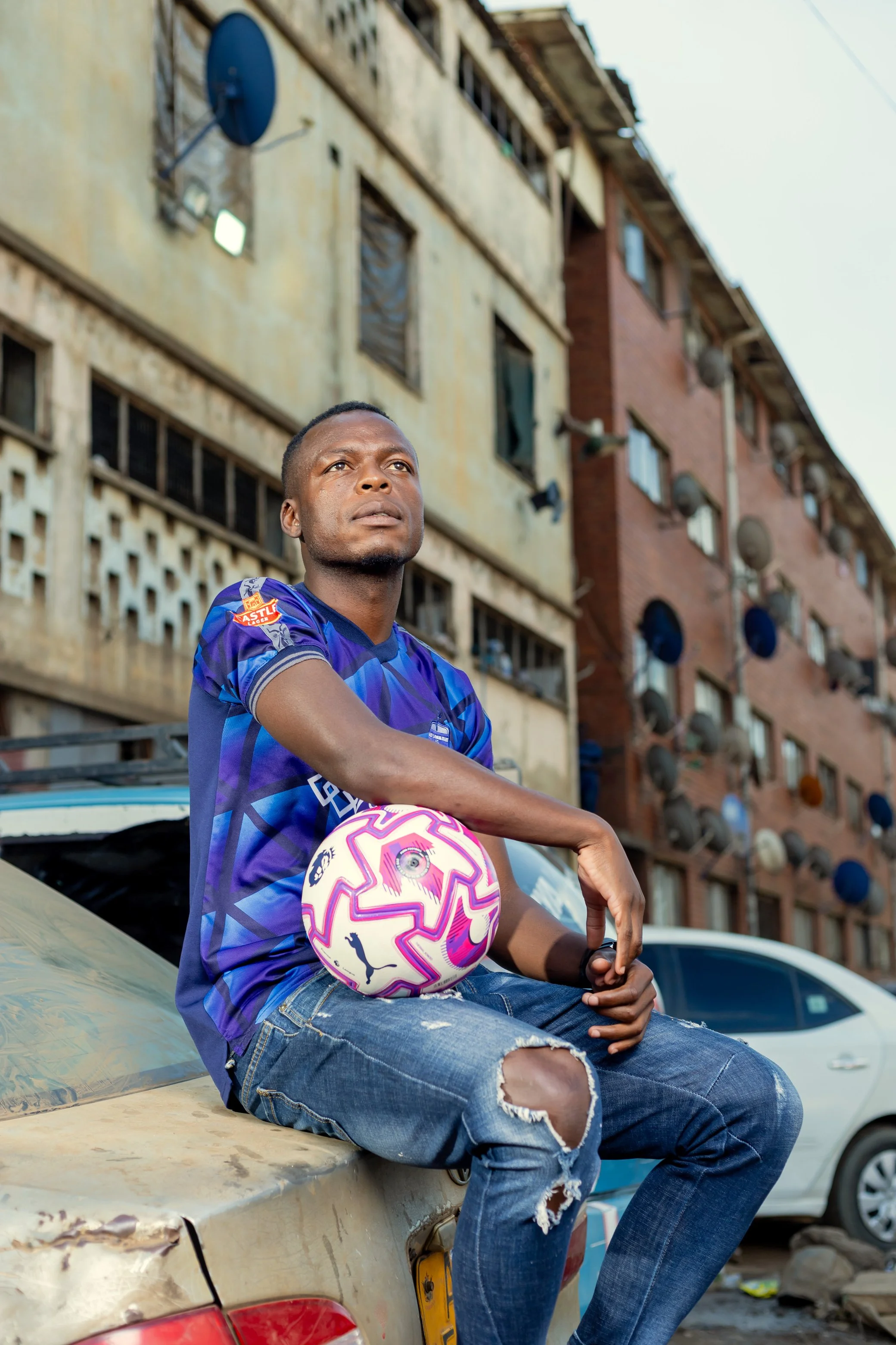 Marvellous Mukumba seated and looking upward while holding a football, photographed against a building in Mbare, Harare