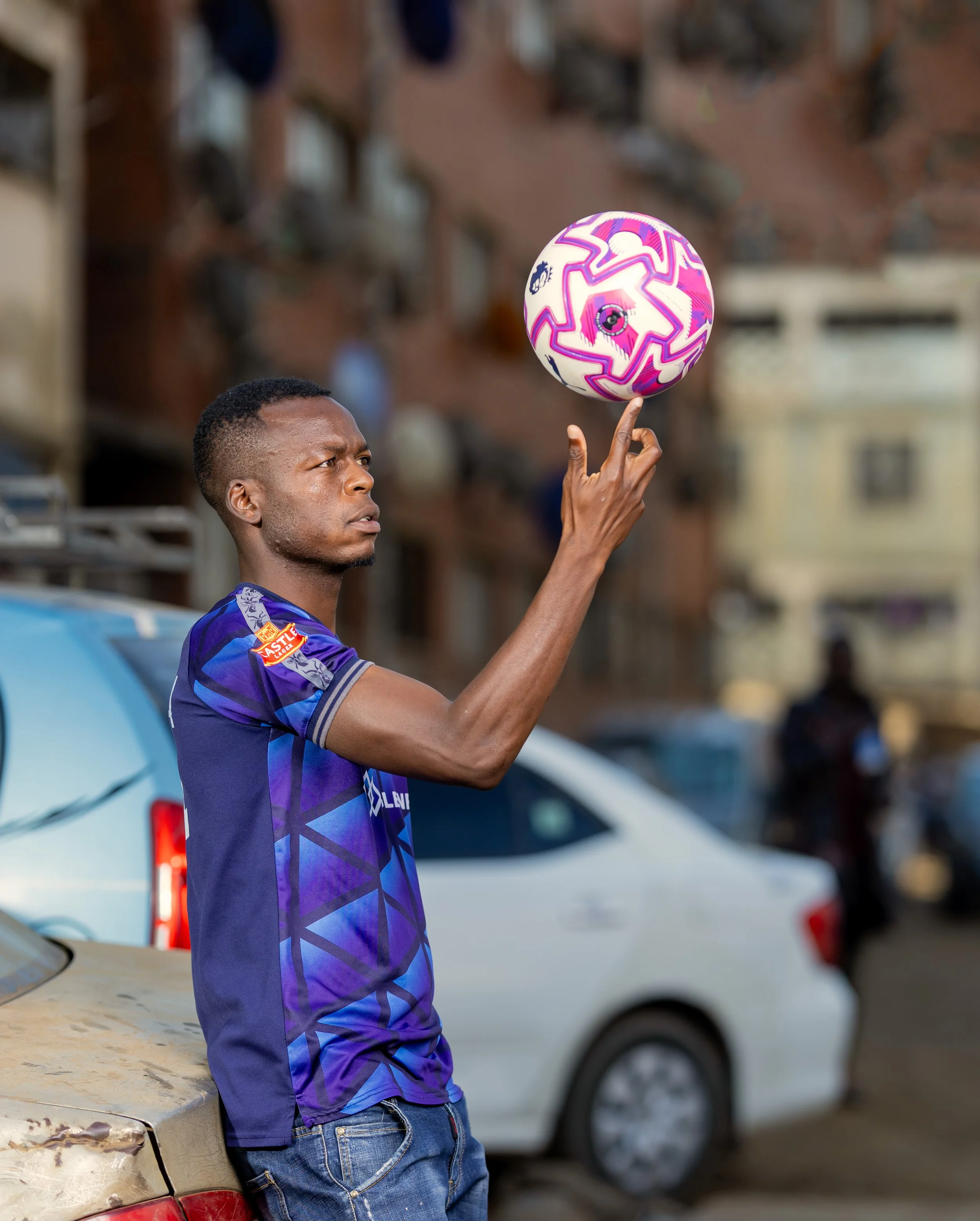Marvellous Mukumba balancing a football on his fingertip on a street in Mbare, Harare, with a parked car and buildings in the background