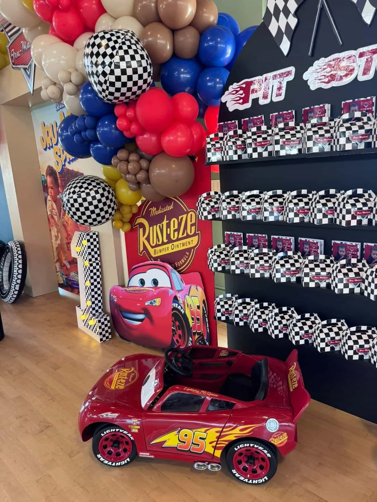 A Disney Pixar Cars themed birthday party setup with a red ride-on car featuring Lightning McQueen, a large number one balloon with a checkered pattern, colorful balloons, and a display of cupcake boxes on a black shelf.