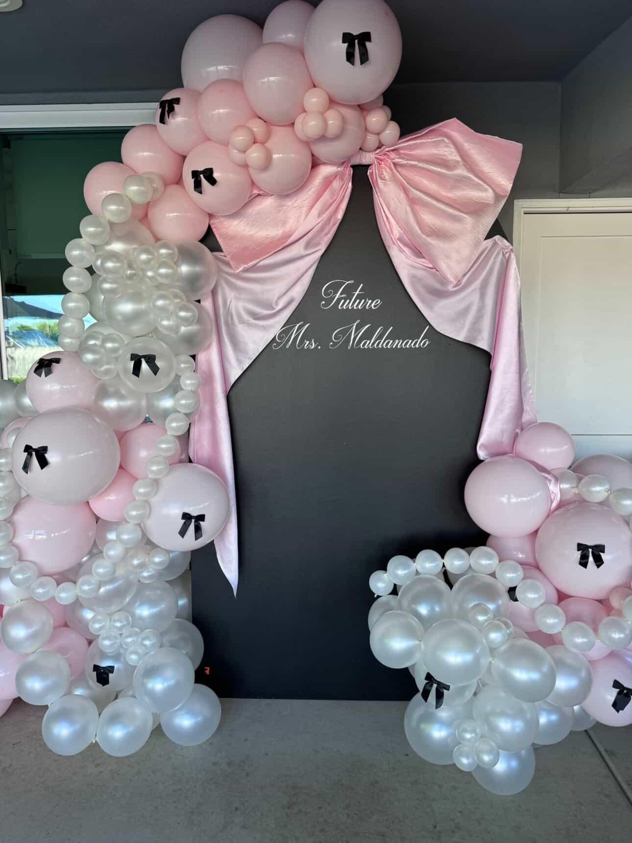 Decorative balloon arch with pink, white, and pearl balloons, featuring small black bows, set against a black backdrop with the white script text "Future Mrs. Maldonado."