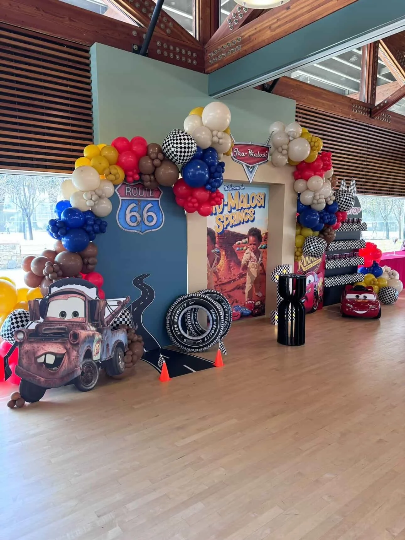 Decorative display with balloons, Cars characters, Route 66 sign, and a large poster for the movie 'Cars' at a party or event. The setup includes colorful balloons arranged in arches, themed cardboard cutouts, and racing accessories.