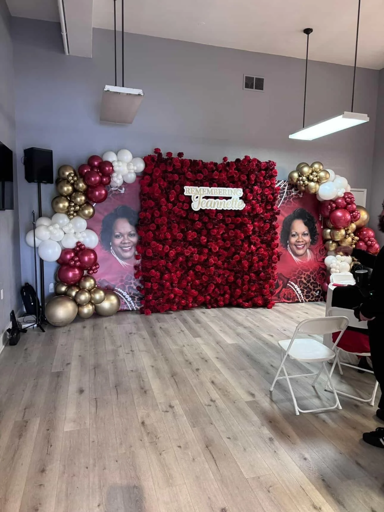 A memorial display with a backdrop of red roses, two large portraits of a smiling woman, gold, white, and maroon balloon arrangements, and a sign reading "Remembering Jeannette" at the top center. The setting appears to be indoors with wooden floorin