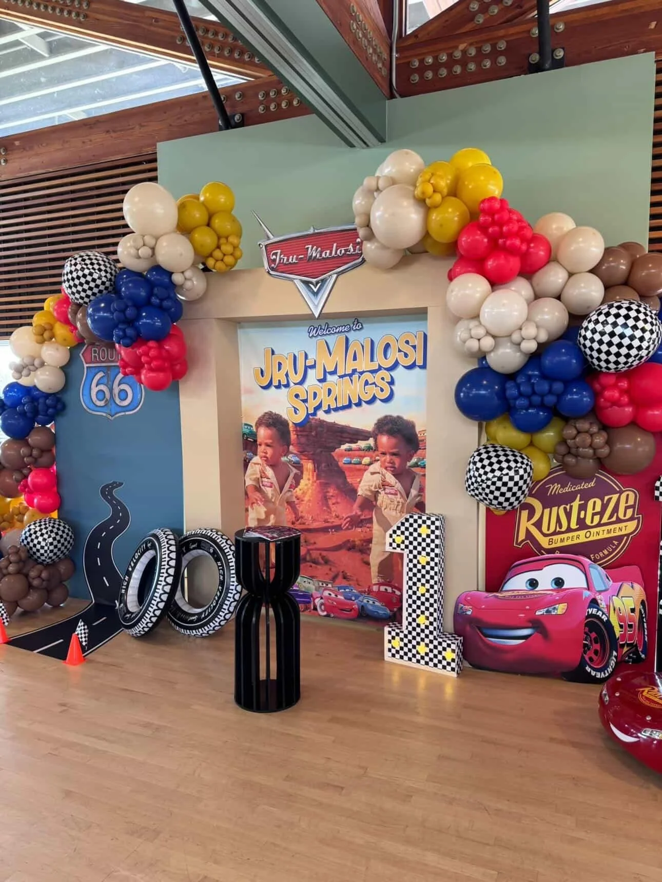 Decorations for a child's birthday party themed for Disney's Cars, featuring balloons, a Cars character cutout, and a Cars-themed backdrop with a poster of children and a desert landscape.