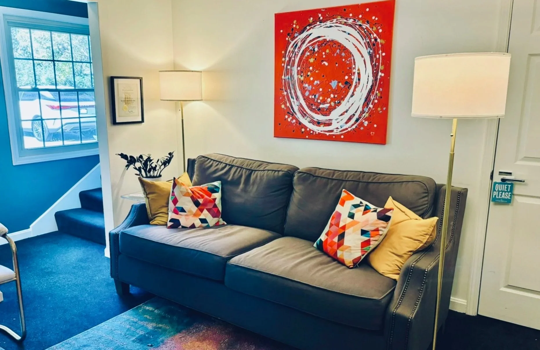 Living room with a grey couch decorated with colorful geometric pillows, a white lamp, a red abstract painting on the wall, a window showing a car outside, and a door with a