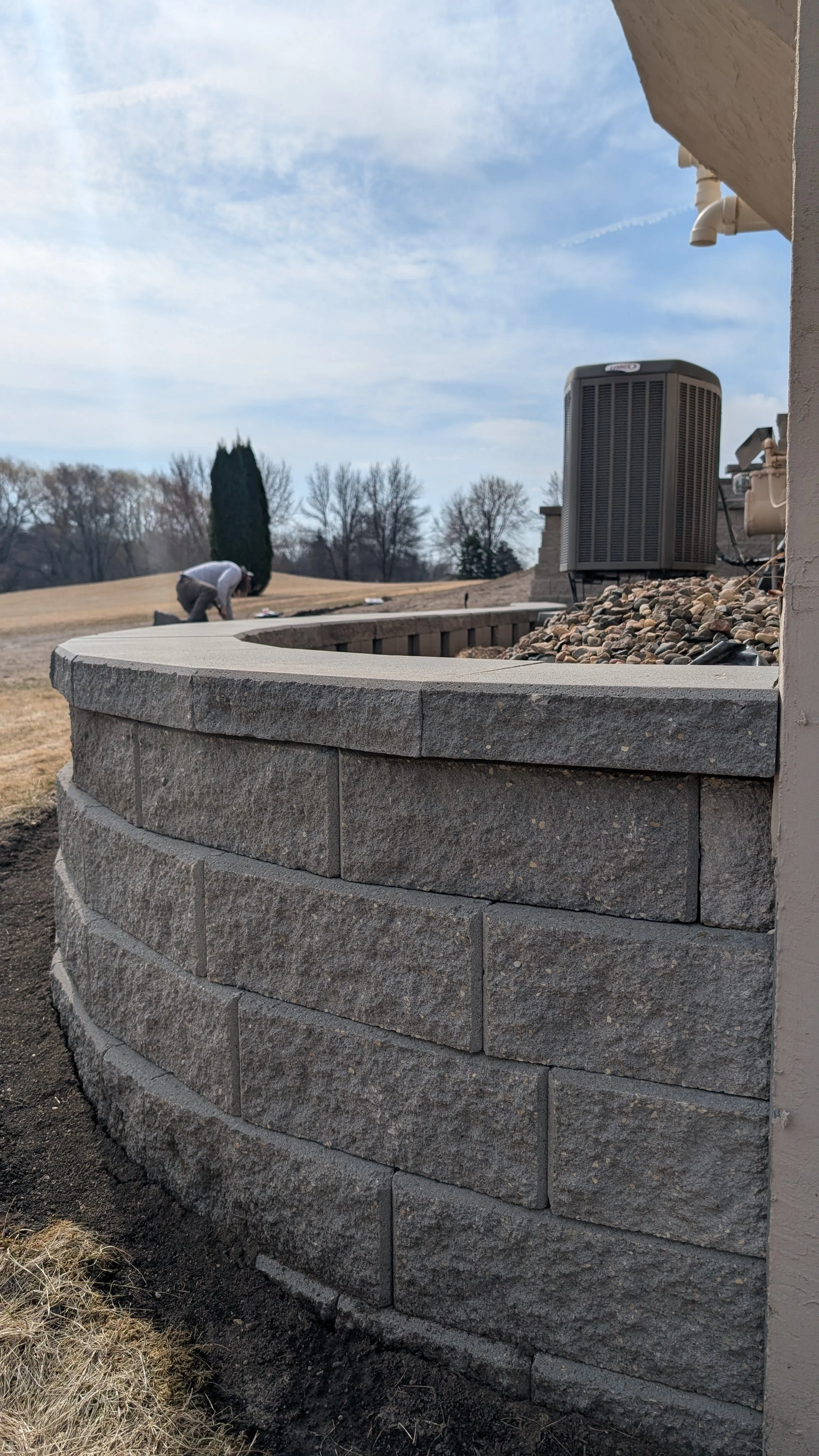 A curved stone retaining wall attached to a building, with an air conditioning unit on top, alongside a landscaped yard with a worker kneeling in the distance.