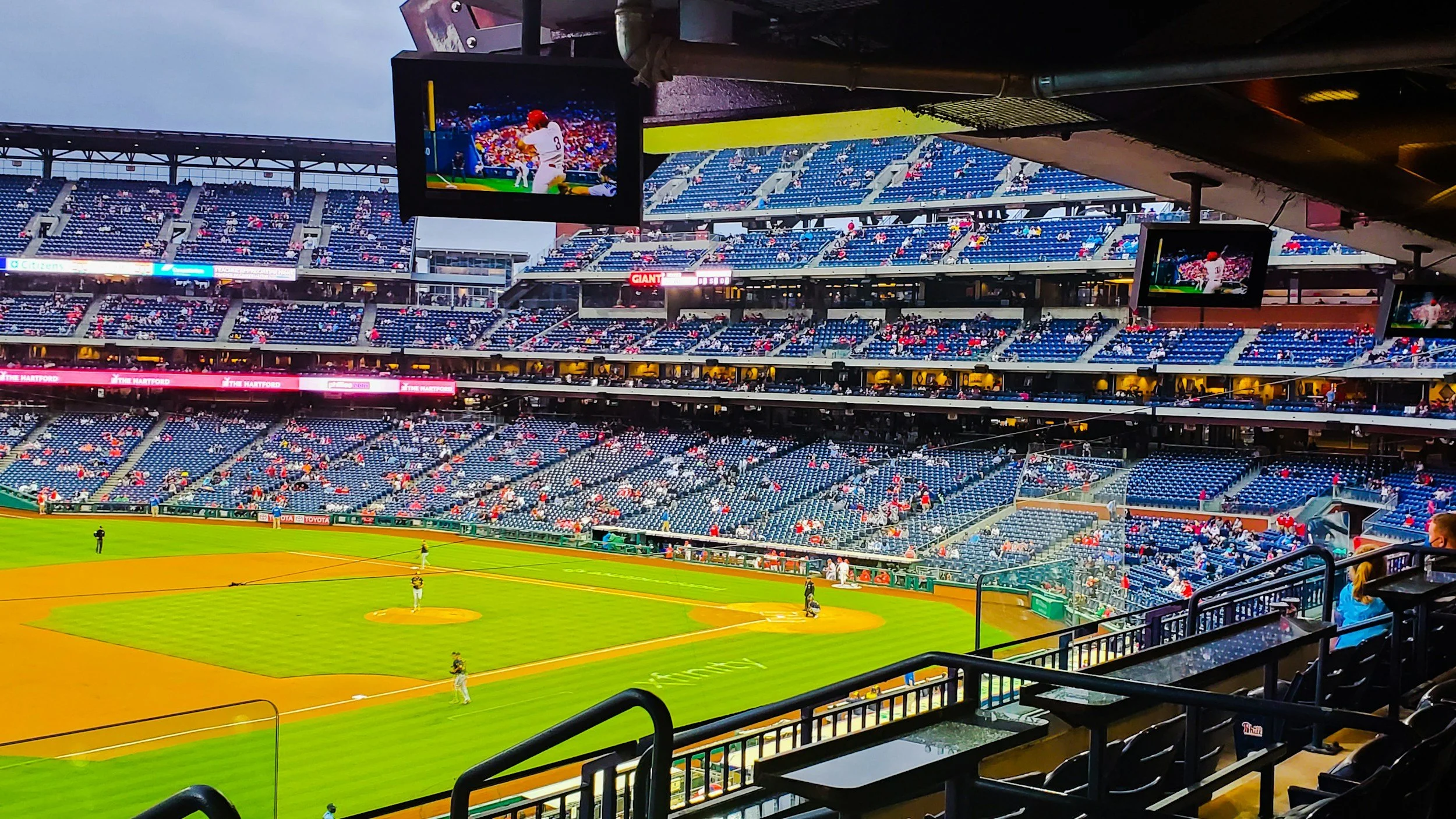 View of a baseball stadium from the seating area, showing the field with players warming up and the empty blue seats in the stands. Multiple screens display the game.