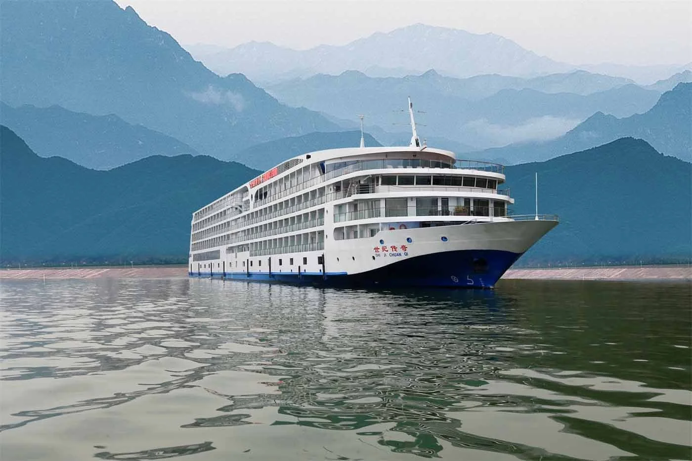 A large white cruise ship docked by a pier on a calm lake, with blue mountains and fog in the background.