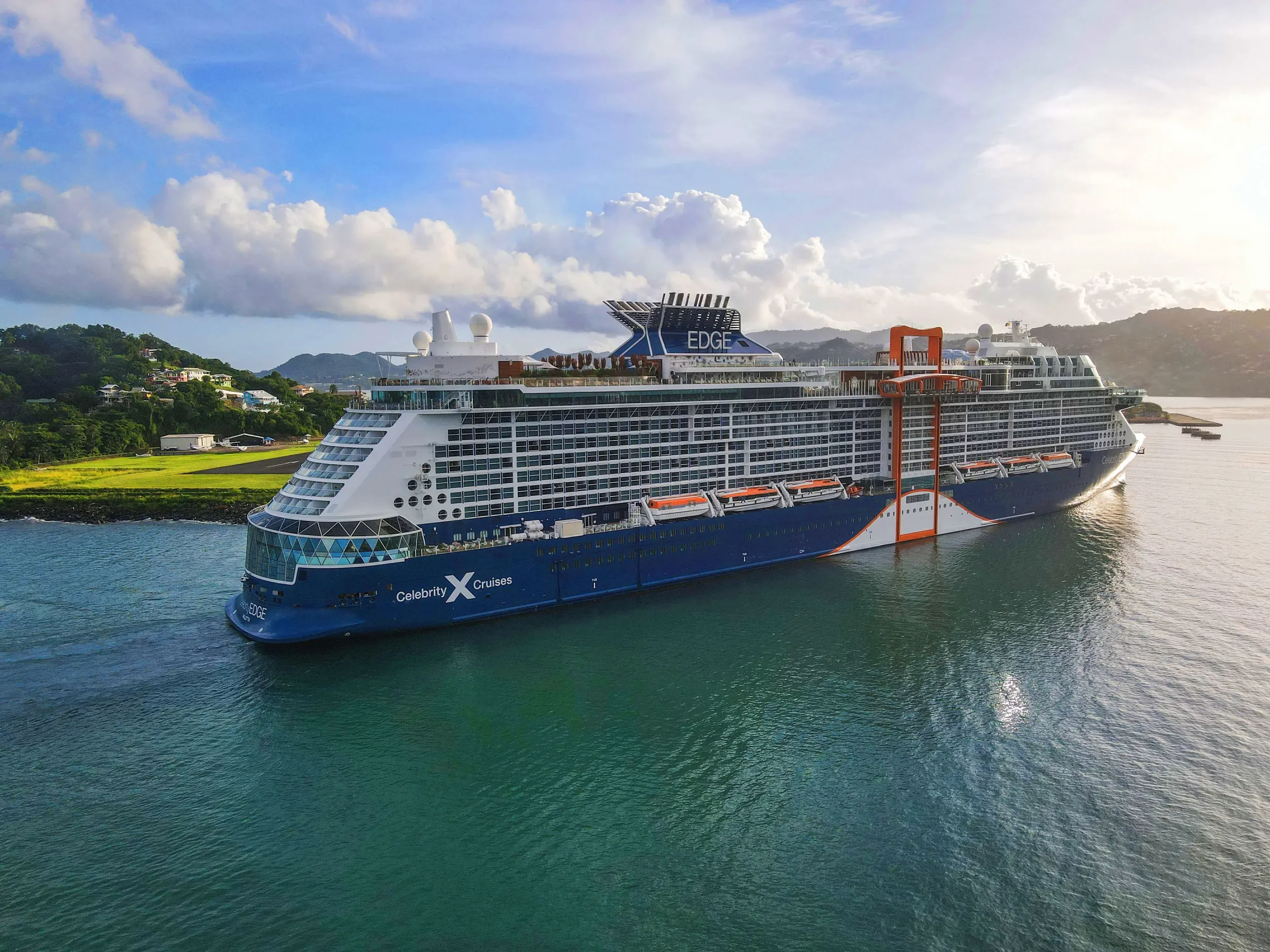 A large cruise ship sailing in a body of water with land and hills in the background, under a partly cloudy sky.