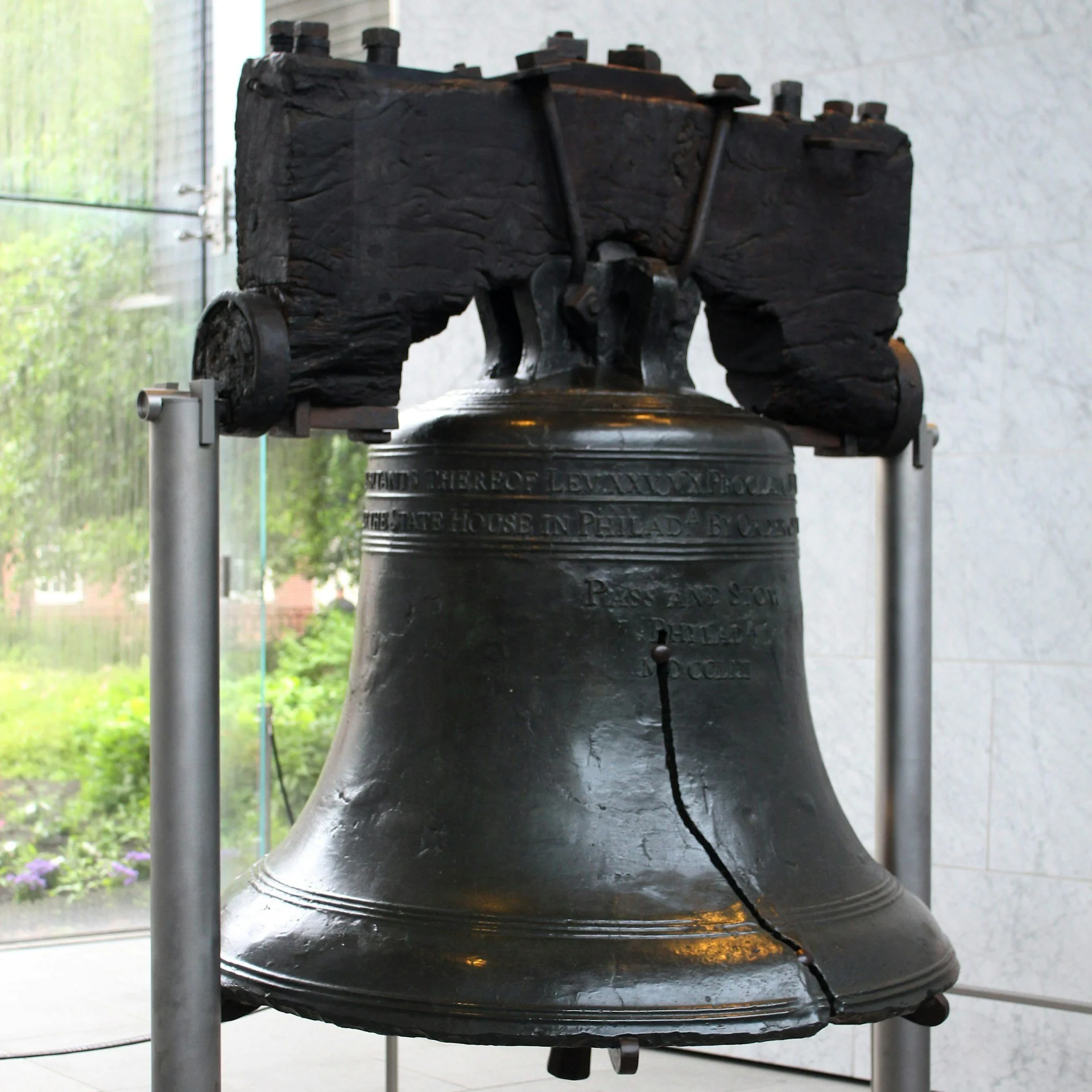 The Liberty Bell displayed indoors with its crack visible, supported by a metal framework, and a window with greenery outside in the background.
