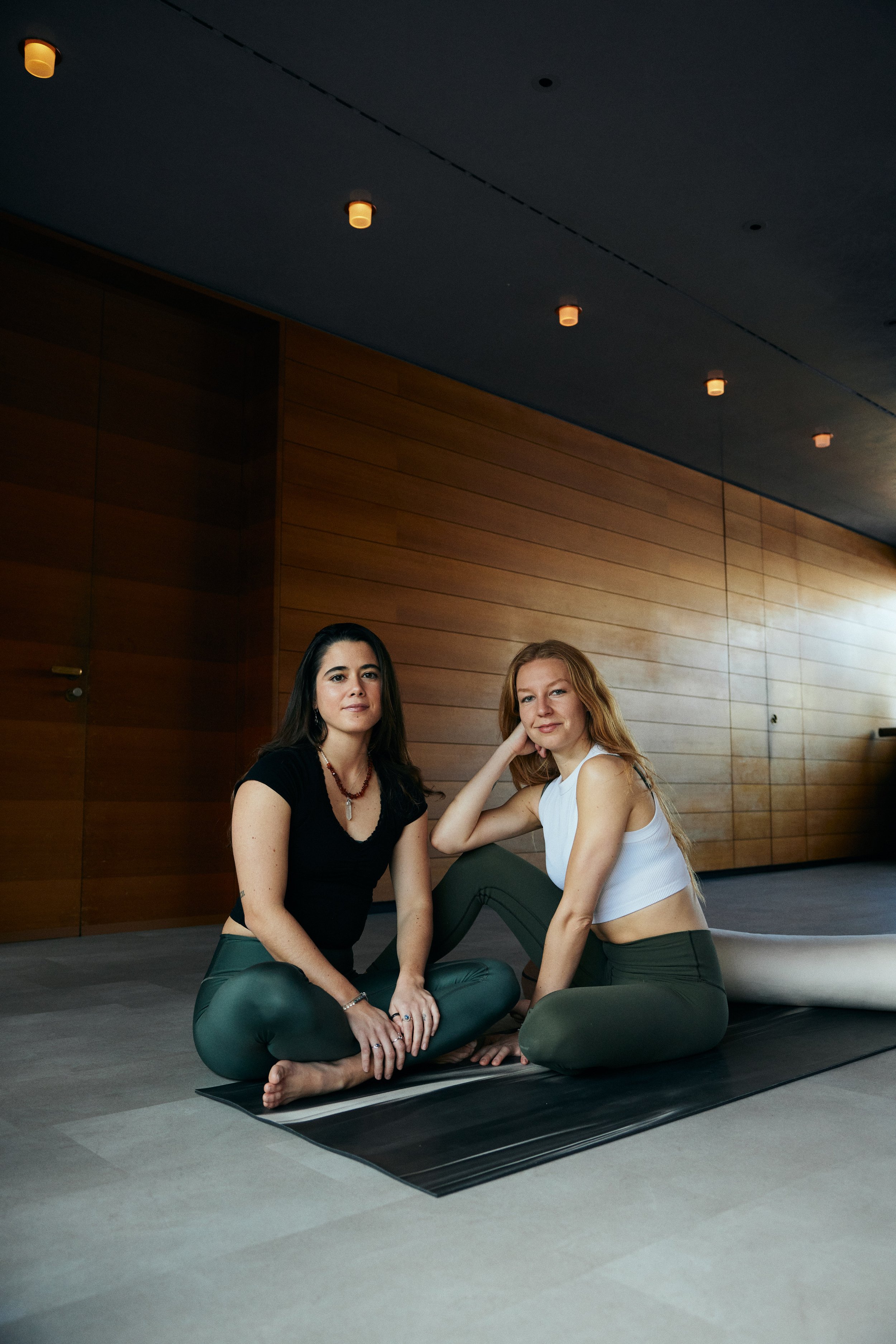 Zwei Frauen sitzen auf einer Yogamatte im Innenraum, vor einer Holzwand, eine trägt eine schwarze T-Shirt und grüne Leggings, die andere ein weißes Top und ebenfalls grüne Leggings.