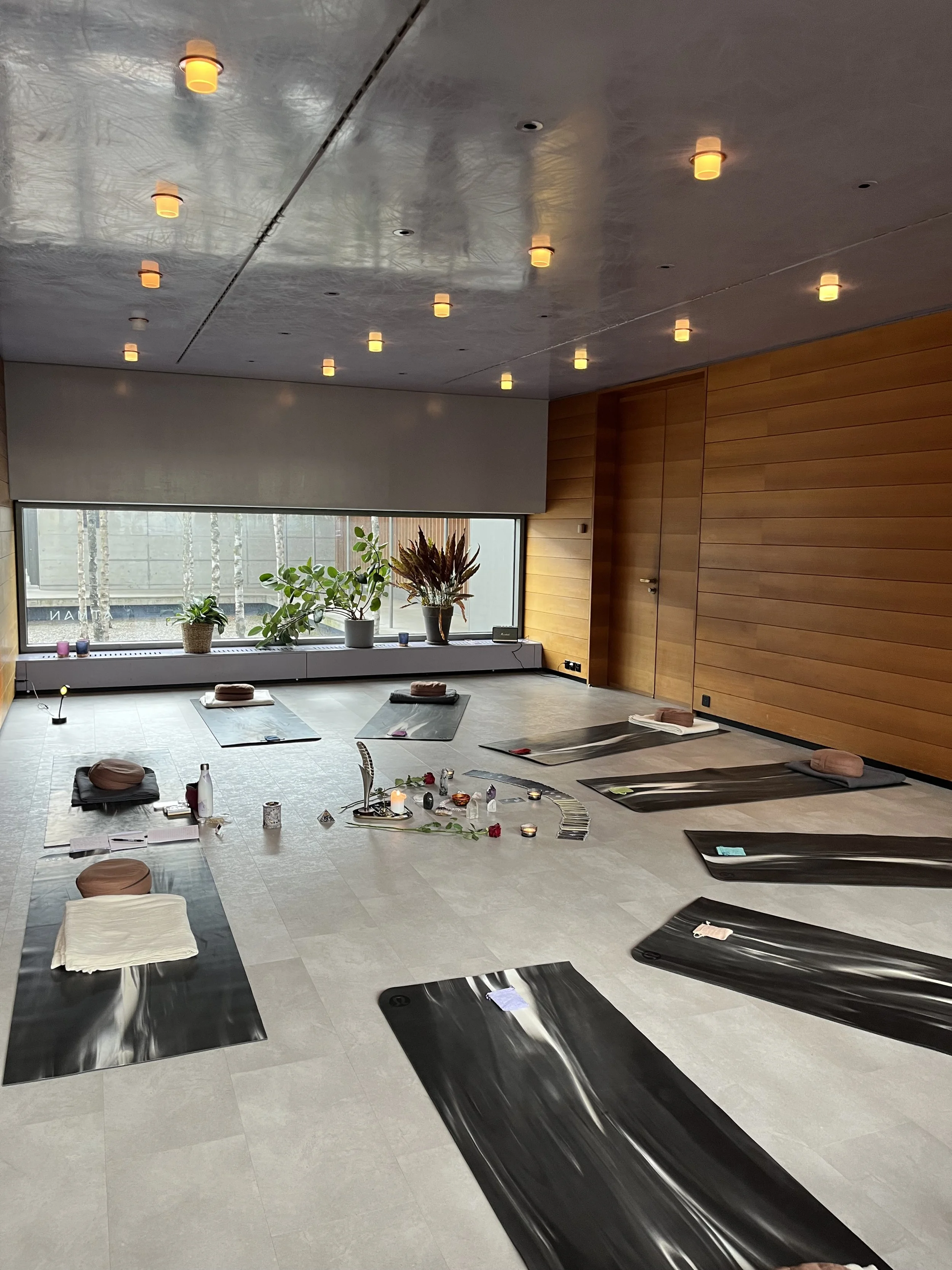 Innenraum eines Yoga- oder Meditationsraums mit Yogamatten, Kissen, Kerzen und Dekorationen, natürliche Tageslicht durch große Fenster, Holzpanelen an den Wänden.