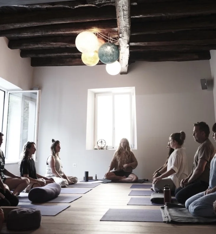 Menschen sitzen im Schneidersitz in einem Raum mit Holzbalkendecke und großen Fenstern, während eine Person in der Mitte einen Meditation oder Yoga Kurs anleitet.