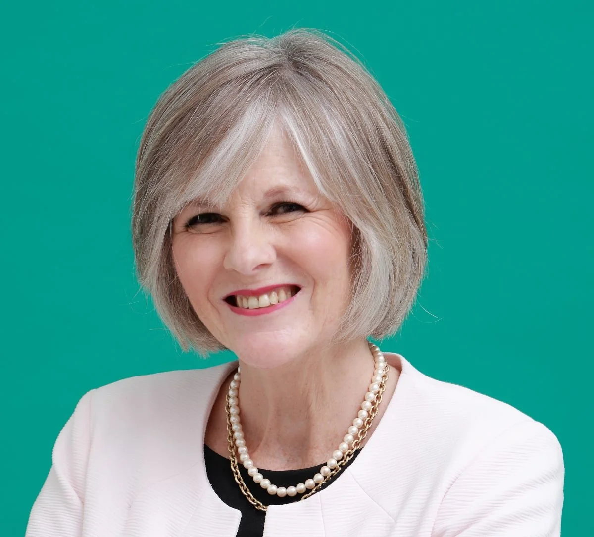 Senior woman with short gray hair smiling, wearing pearl necklace and white top, against a teal background.