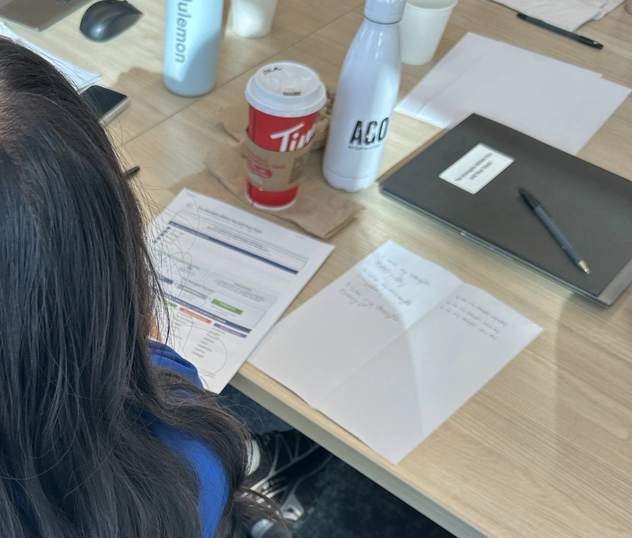 Overhead view of a work desk with papers, a pen, a closed laptop, beverages, and a person with long dark hair sitting nearby.