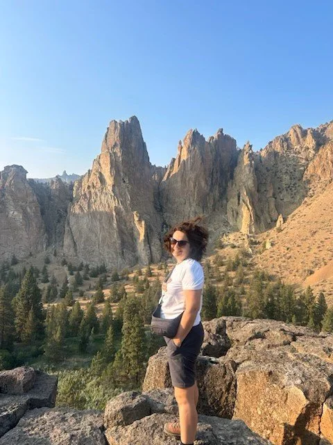 Woman wearing sunglasses, white t-shirt, and black shorts standing on rocks with rocky mountain peaks and pine trees in the background during daytime.
