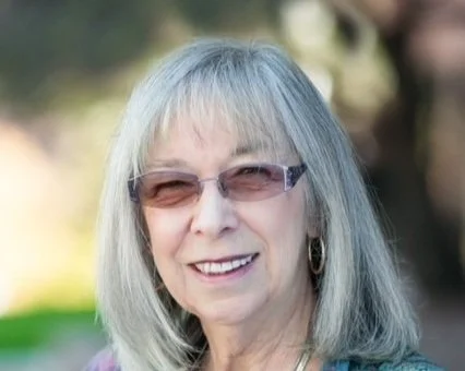 A smiling older woman with shoulder-length gray hair, wearing sunglasses and hoop earrings, outdoors with blurred greenery in the background.
