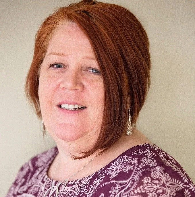 A woman with short red hair, wearing a purple patterned blouse and dangling earrings, smiling at the camera against a plain background.