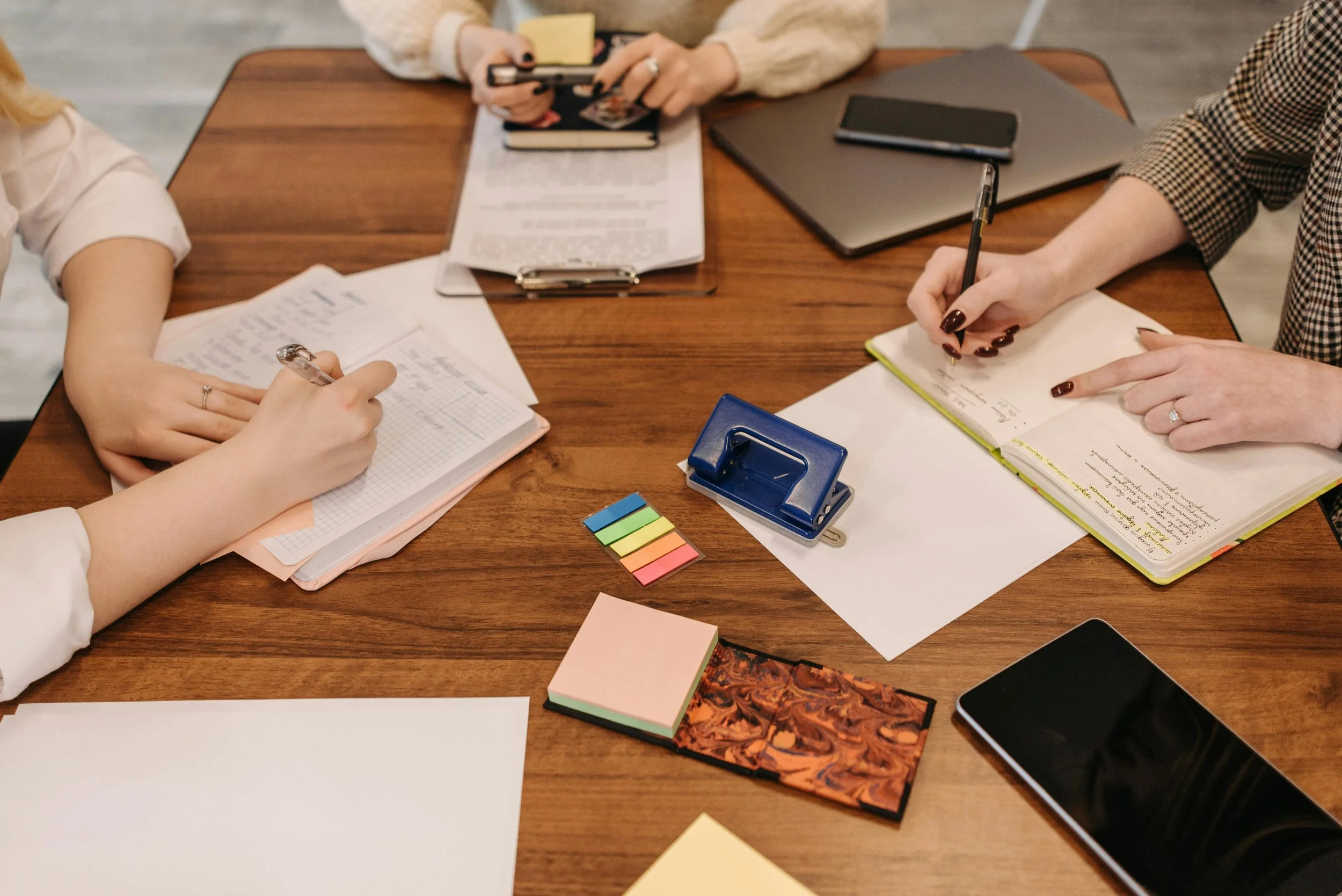 People sitting around a wooden table, writing in notebooks, with various office supplies like pens, sticky notes, and electronic devices on the table.