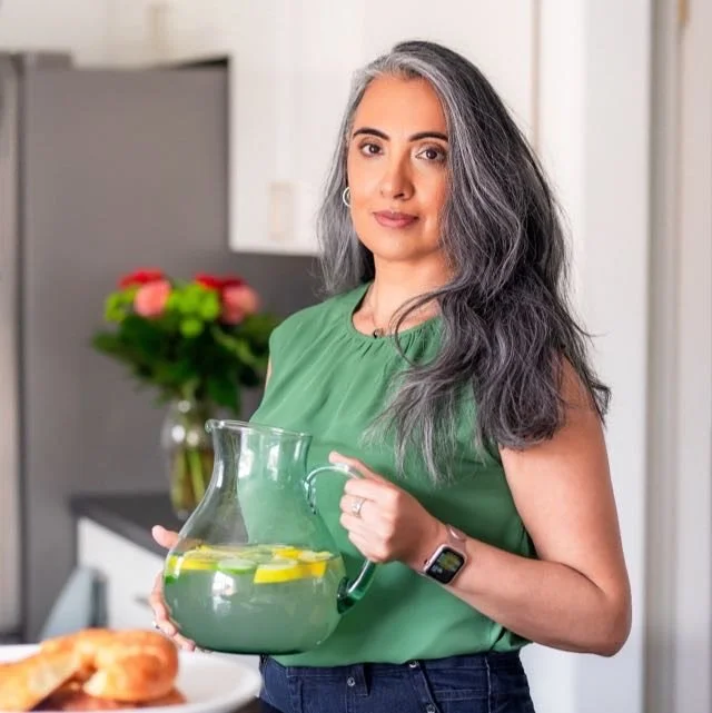 A woman with long gray hair wearing a green sleeveless top and a smartwatch, standing indoors and holding a glass pitcher of lemonade with lemon slices, with a vase of colorful flowers on a kitchen counter in the background.