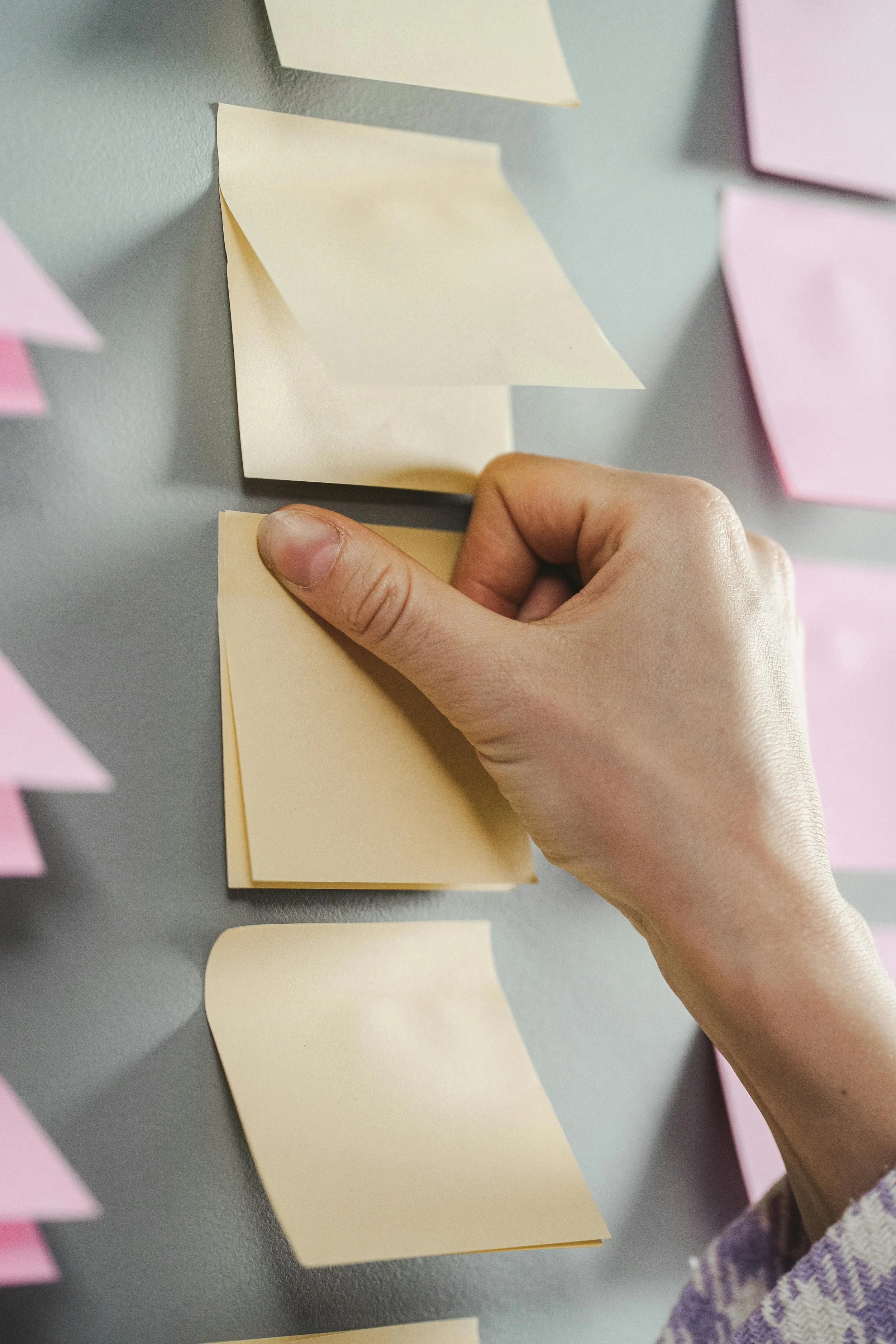 A person's hand placing a yellow sticky note on a greyboard with other yellow and pink sticky notes attached.