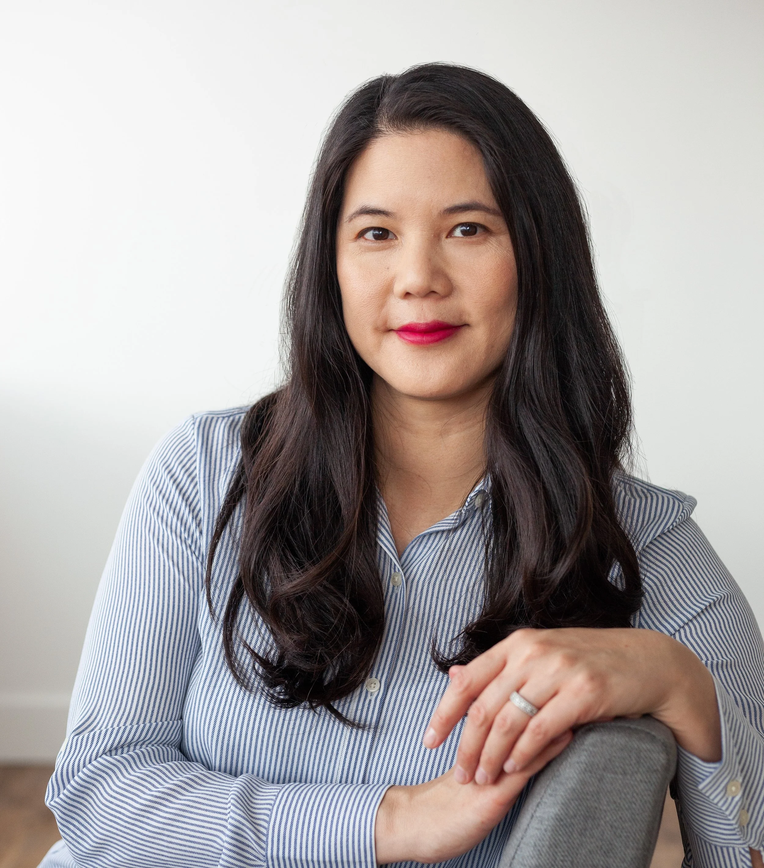 Professional woman with long dark hair wearing a black blazer and top standing against a plain light background, with plants blurred in the background.