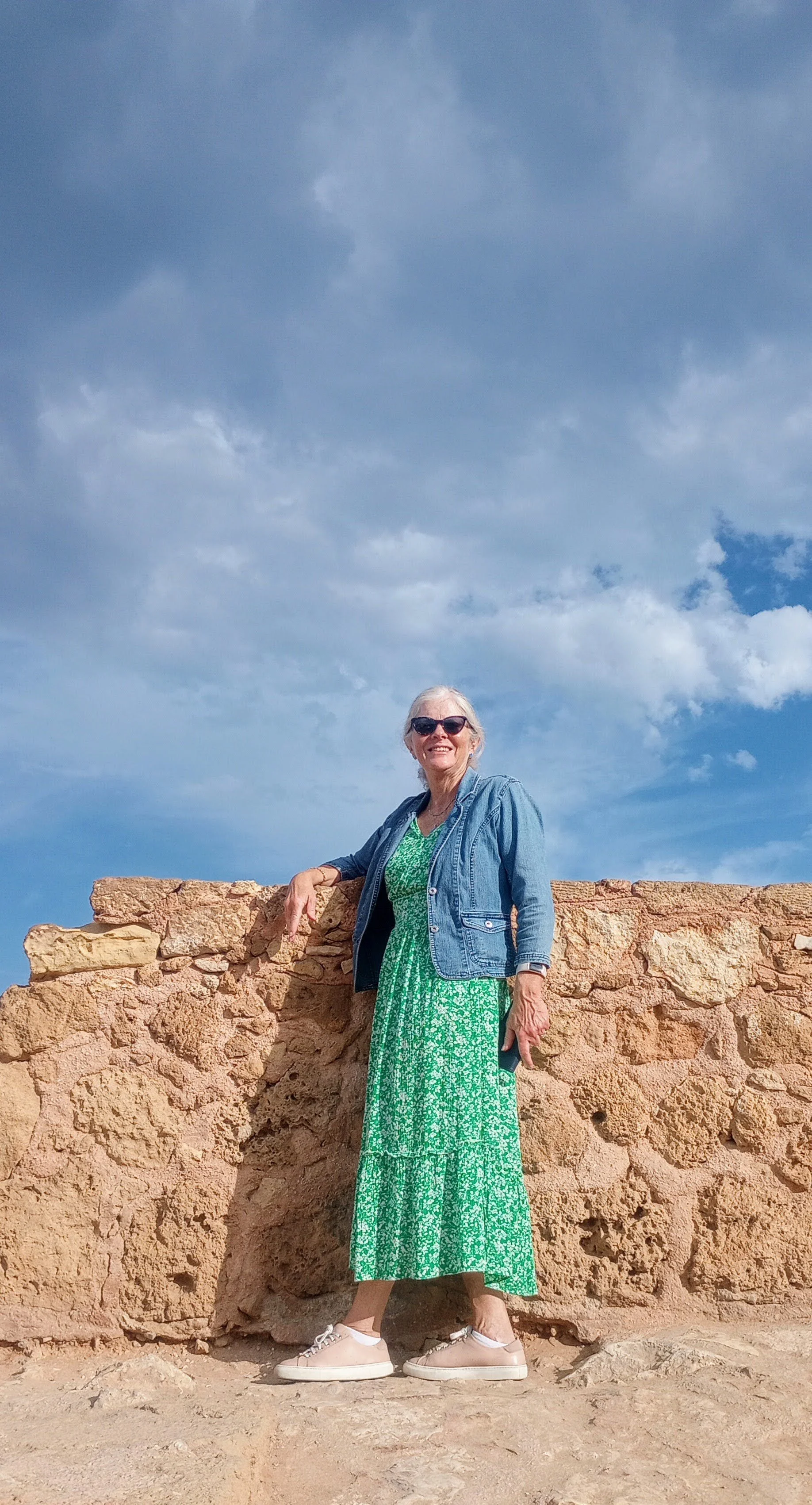 Smiling elderly woman wearing sunglasses, a green floral dress, a denim jacket, and beige sneakers standing outdoors against a stone wall with a blue sky and clouds in the background.