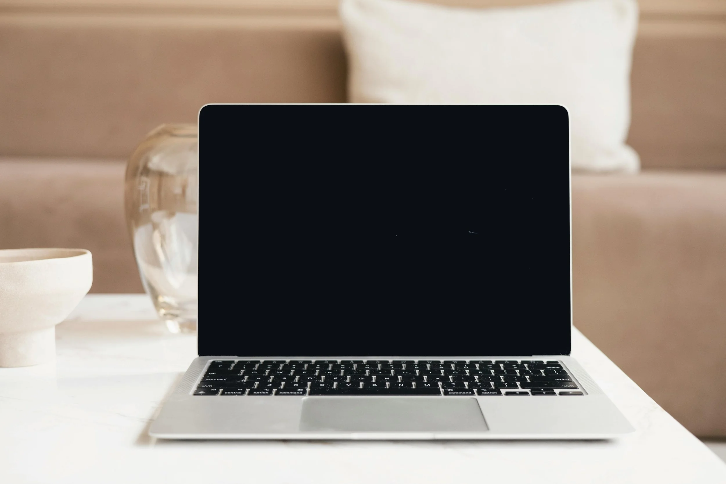 Open silver laptop on a white table with a beige couch, decorative glass vase, and a ceramic bowl in the background.