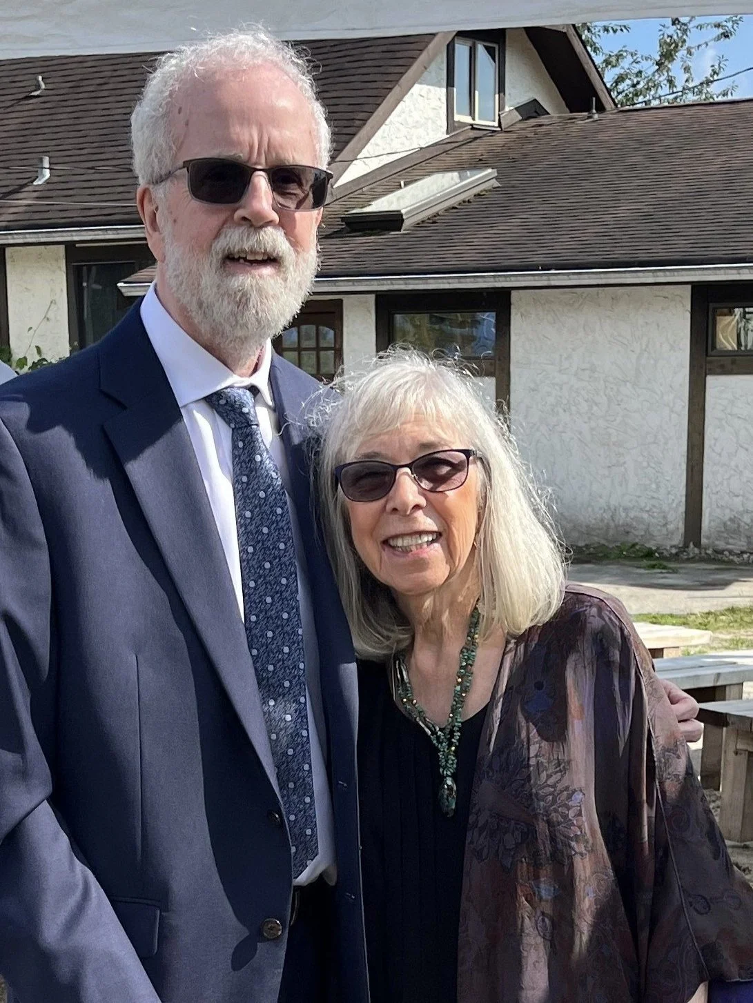 A man and woman wearing sunglasses standing outside in front of a house on a sunny day. The man is dressed in a blue suit with a patterned tie and has white hair and a beard. The woman has shoulder-length blonde hair, is wearing a colorful jacket, a black top, and a necklace.