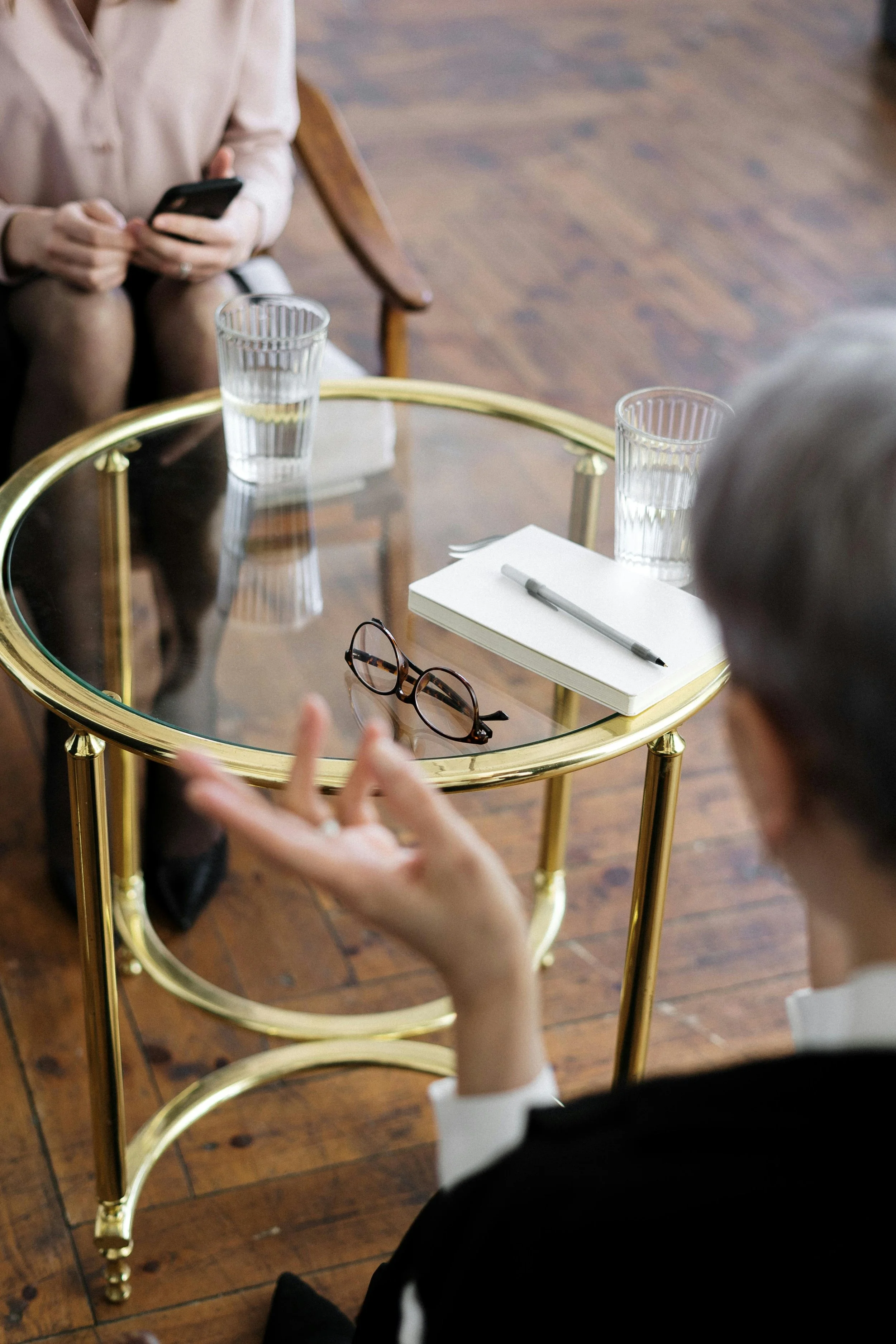 A glass-top gold table with a pair of glasses, a notebook, and a pen on it. Two people are seated around the table, one woman holding a phone, and a man gesturing with his hand.
