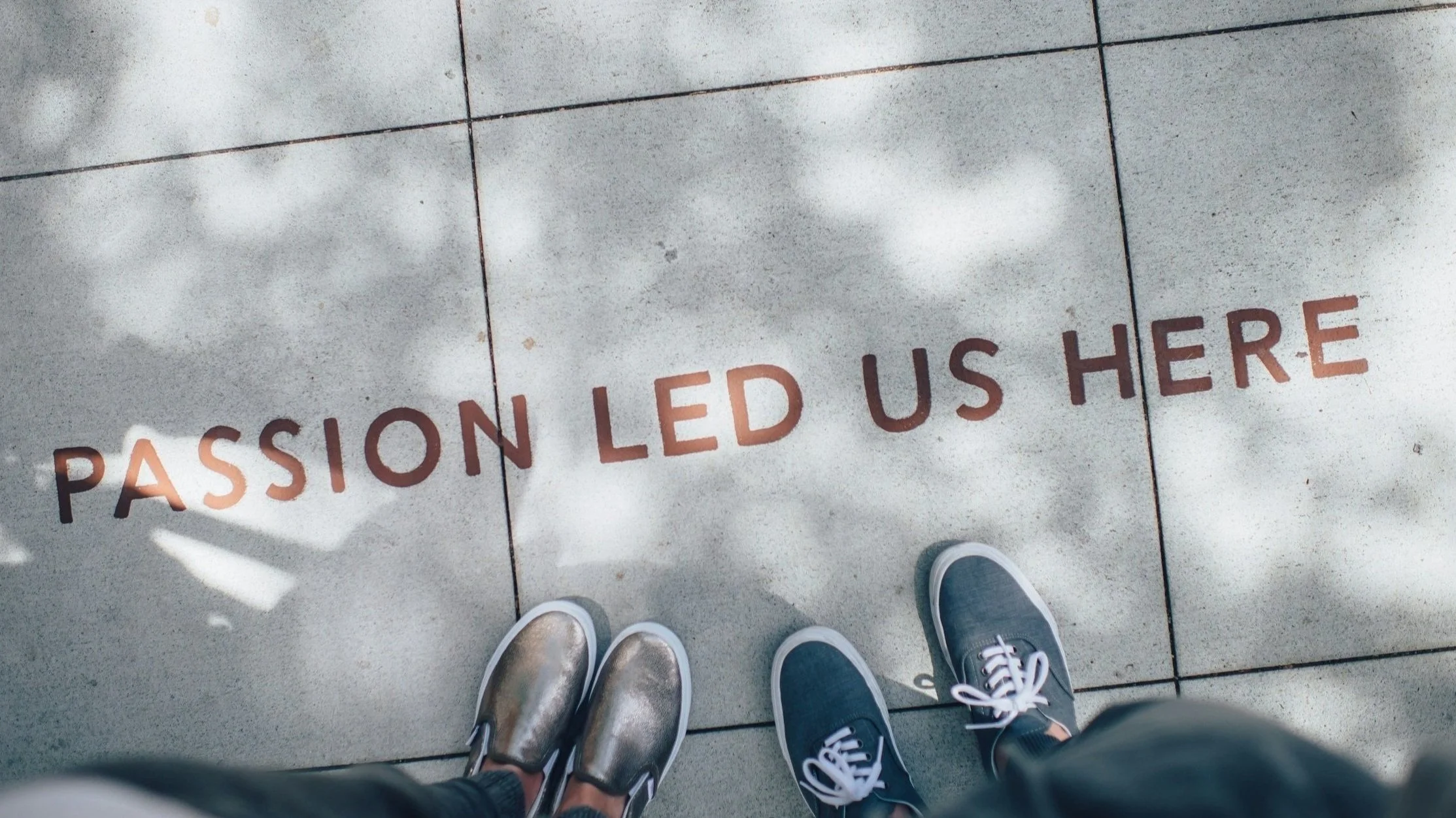 Close-up of two pairs of shoes and a message on the sidewalk that says 'passion led us here', with reflections of clouds on the sidewalk.