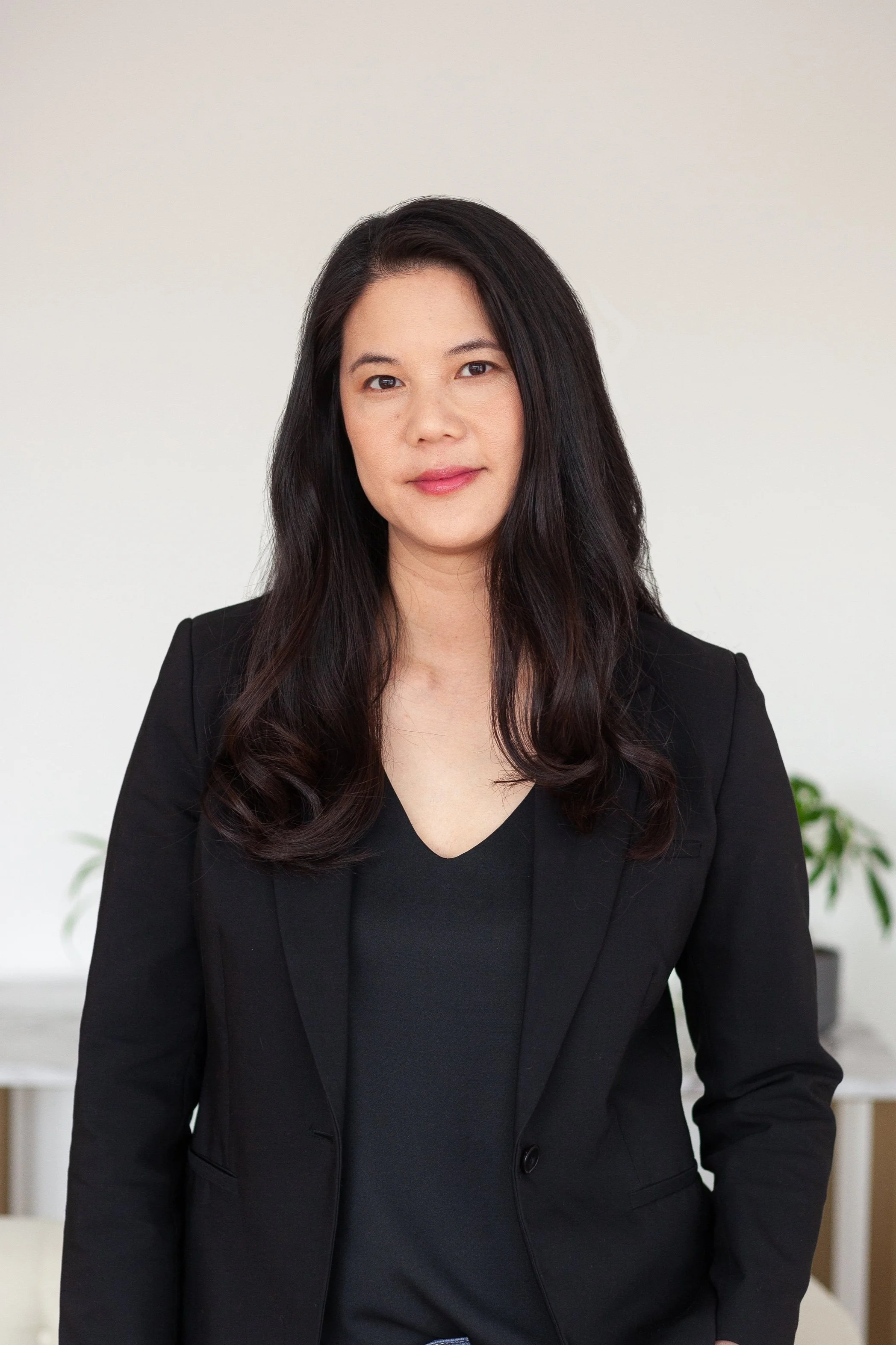 Professional woman with long dark hair wearing a black blazer and top standing against a plain light background, with plants blurred in the background.